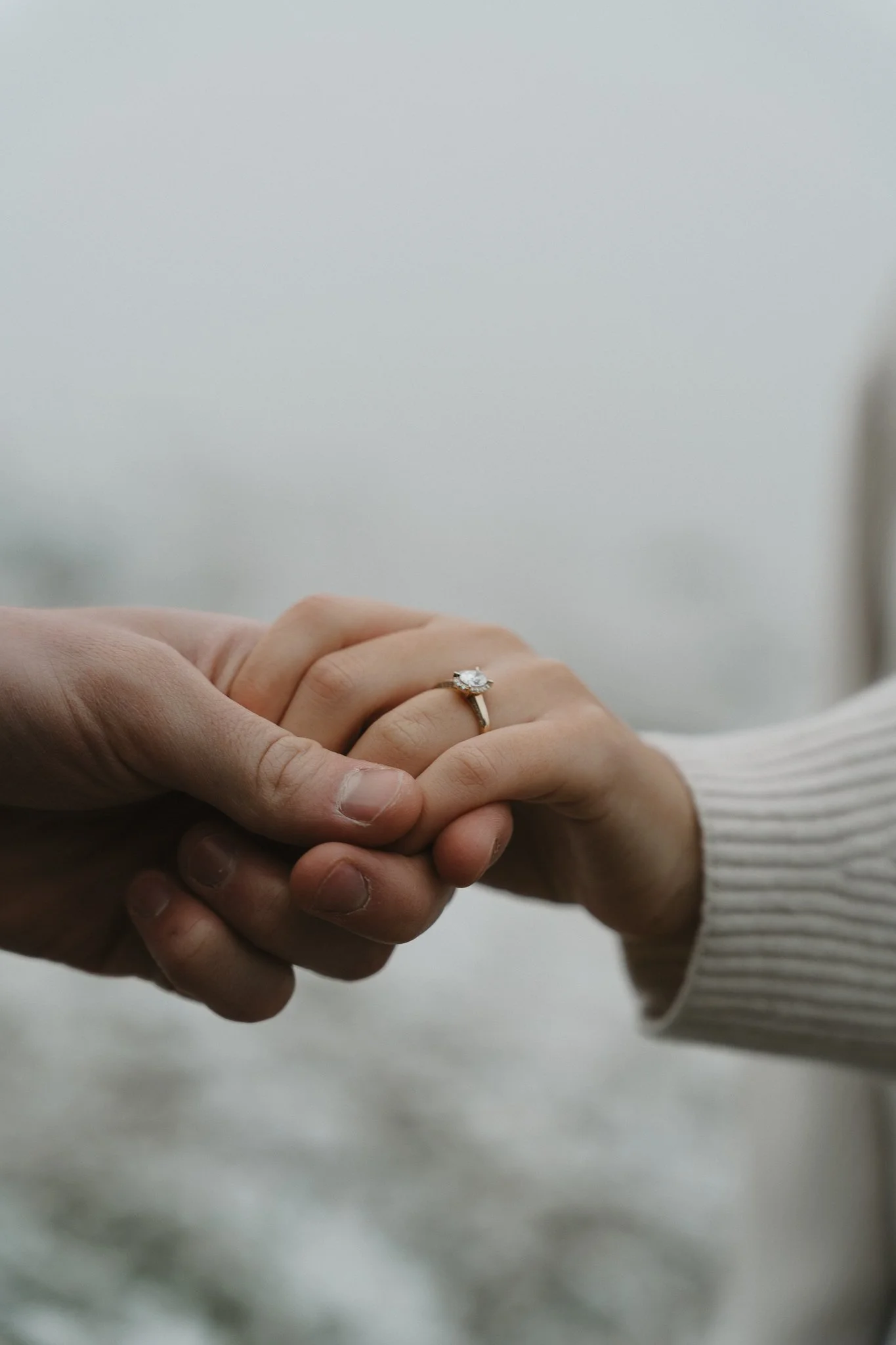Close-up of a person's hand with an engagement ring holding another person's hand, against a foggy background.