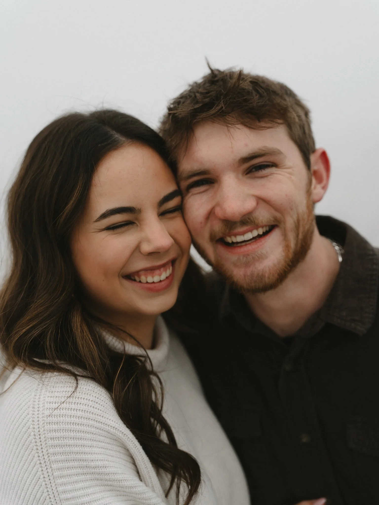 A smiling young woman with long brown hair and a young man with short light brown hair and a beard, both with eyes closed, happy, close together, against a plain white background.