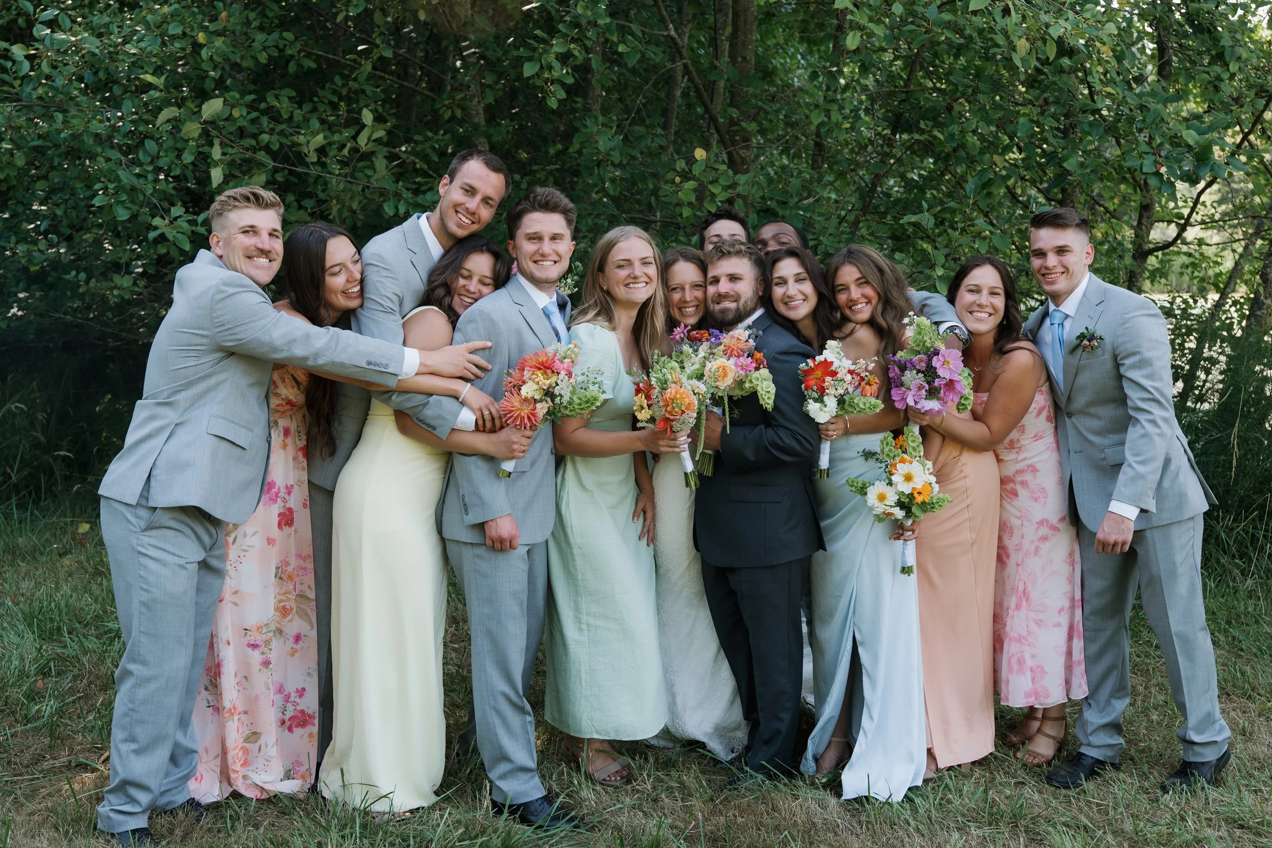 A wedding party of 15 people posing outdoors with trees in the background, dressed in formal attire, holding colorful bouquets of flowers, smiling, celebrating the wedding.