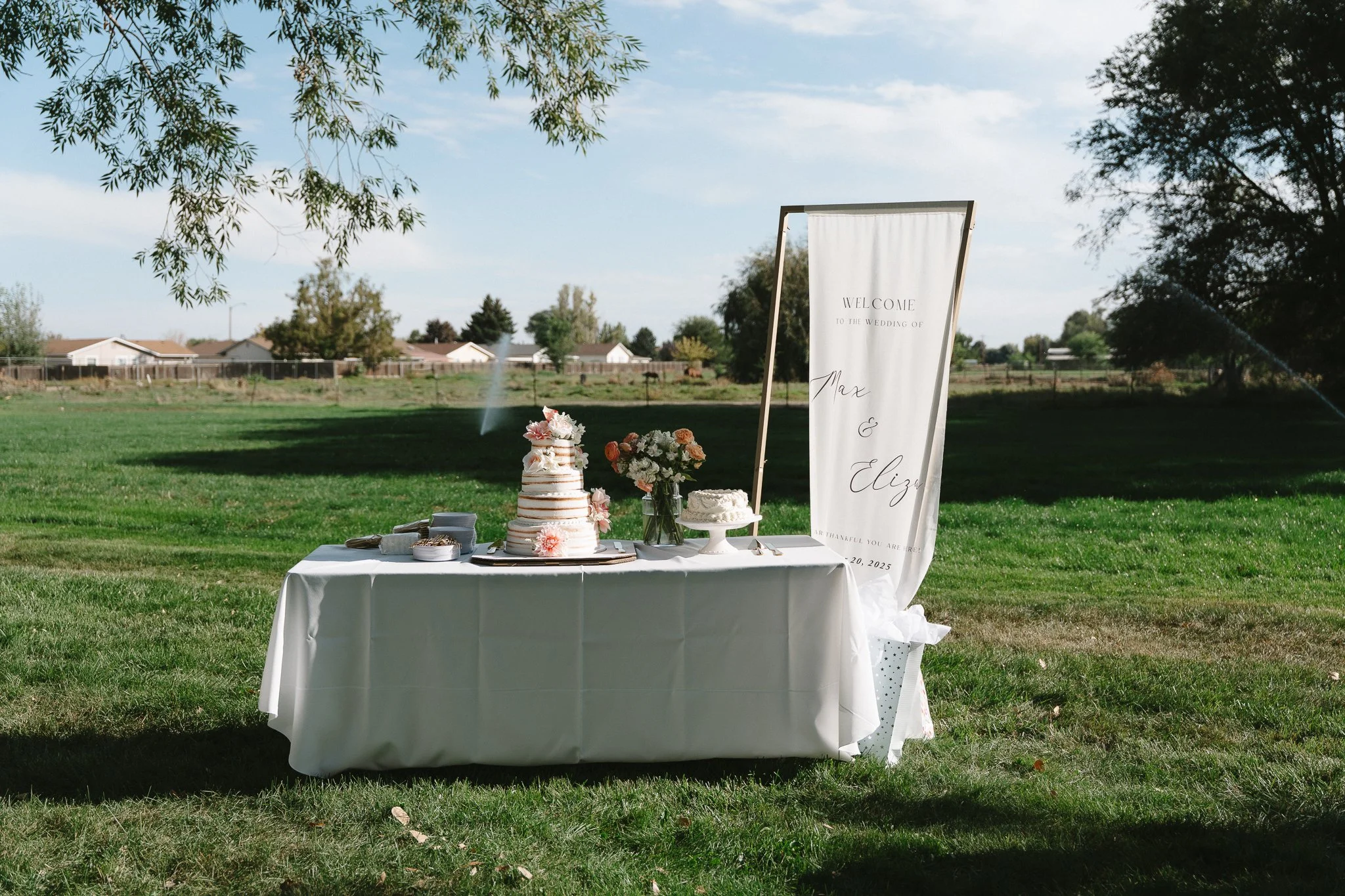 Outdoor wedding reception table with a wedding cake, floral arrangements, and a welcoming sign on a grassy field with trees and houses in the background.