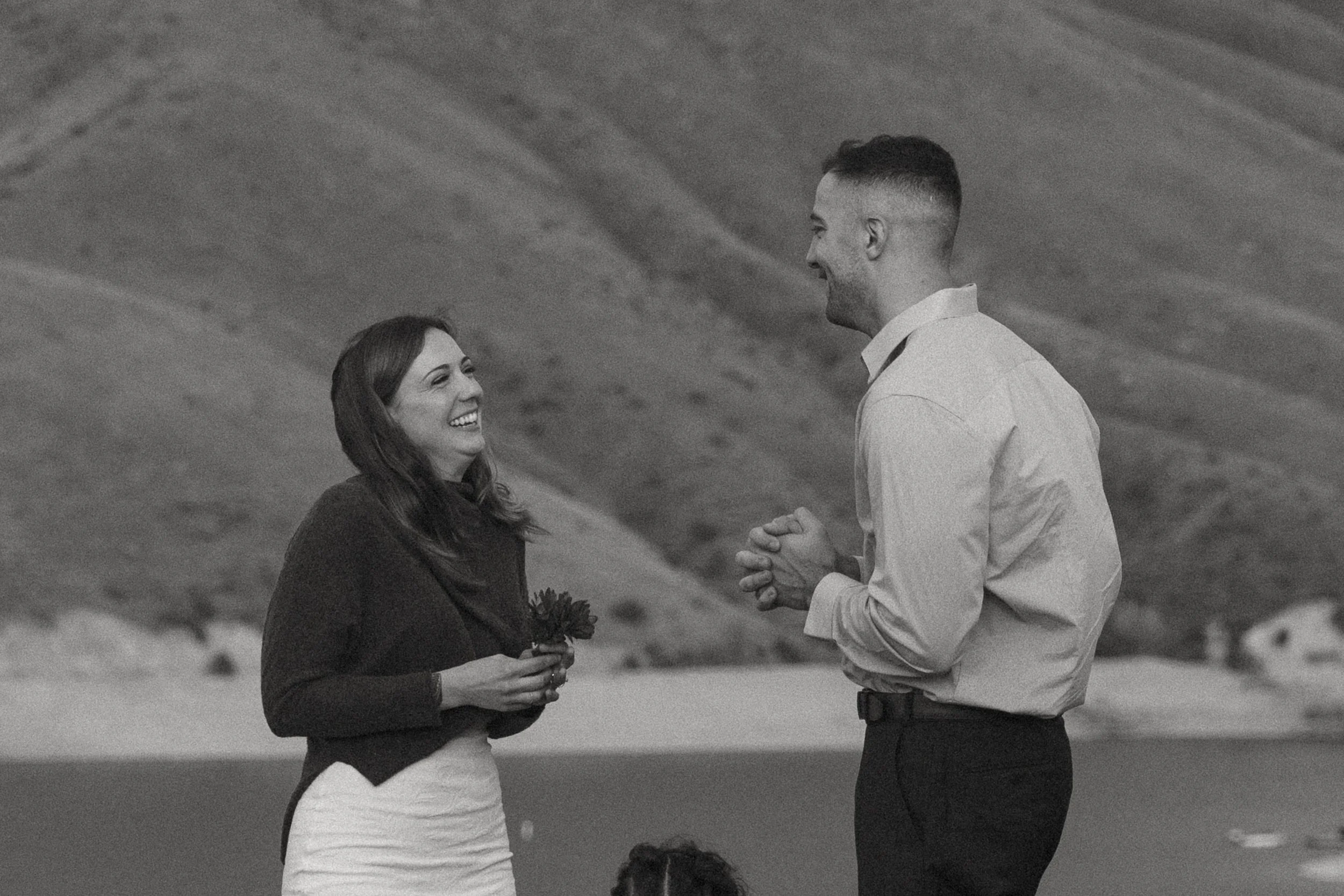 A woman and a man are smiling and holding hands, standing outdoors on a slight incline with a hilly landscape in the background.