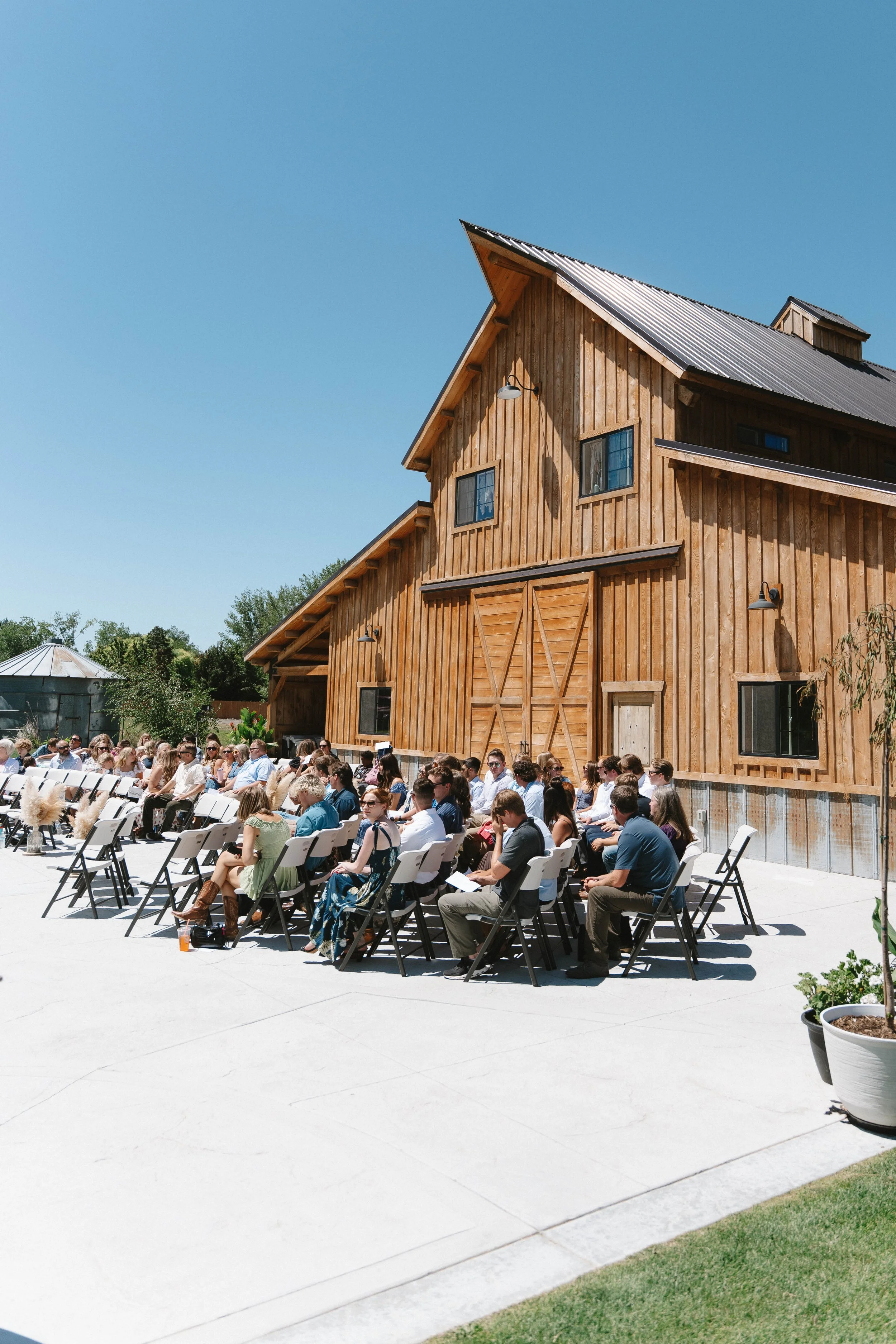 People seated outdoors near a large wooden barn during a sunny day.