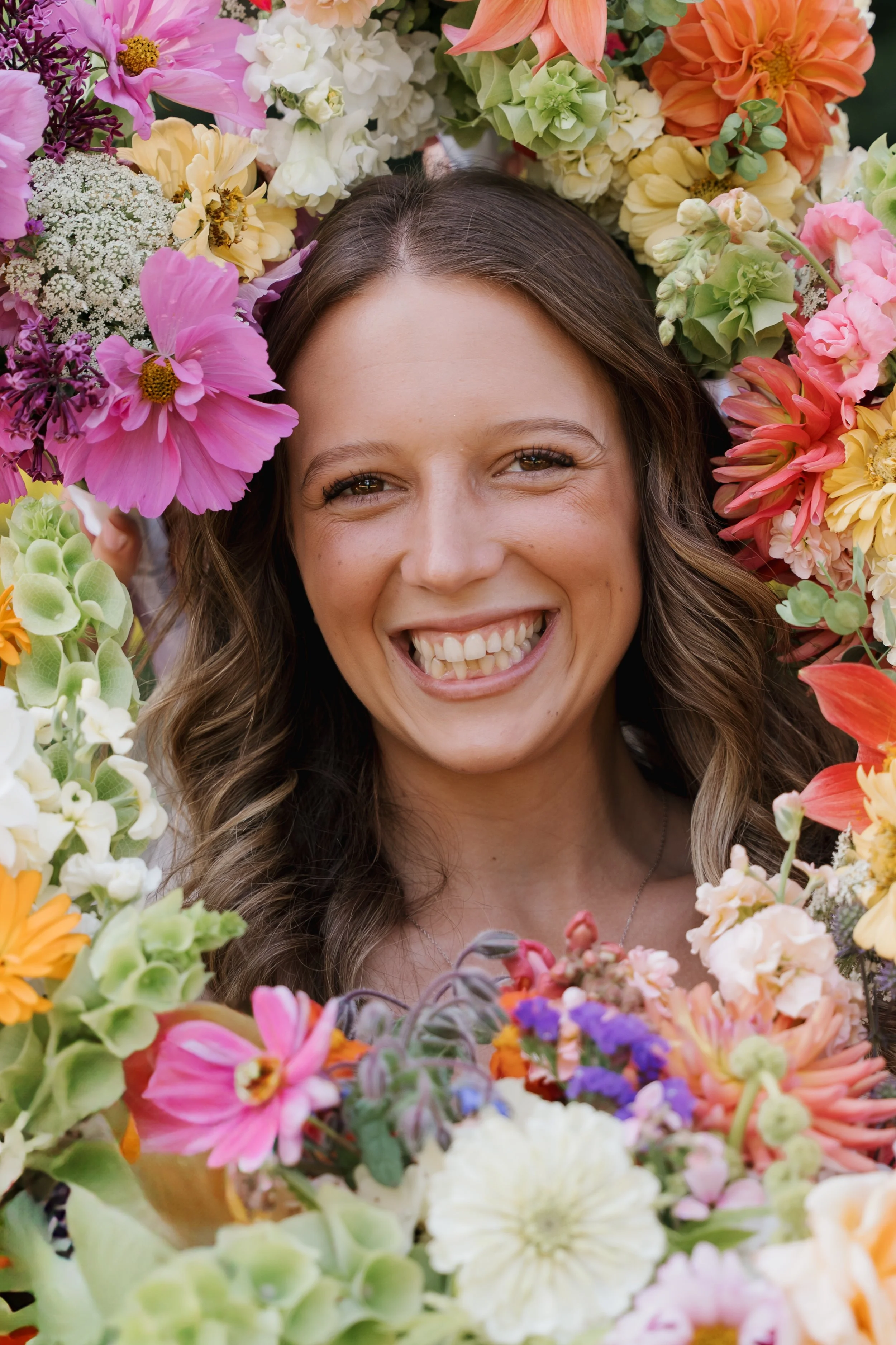 A smiling woman with brown hair surrounded by an assortment of colorful flowers.