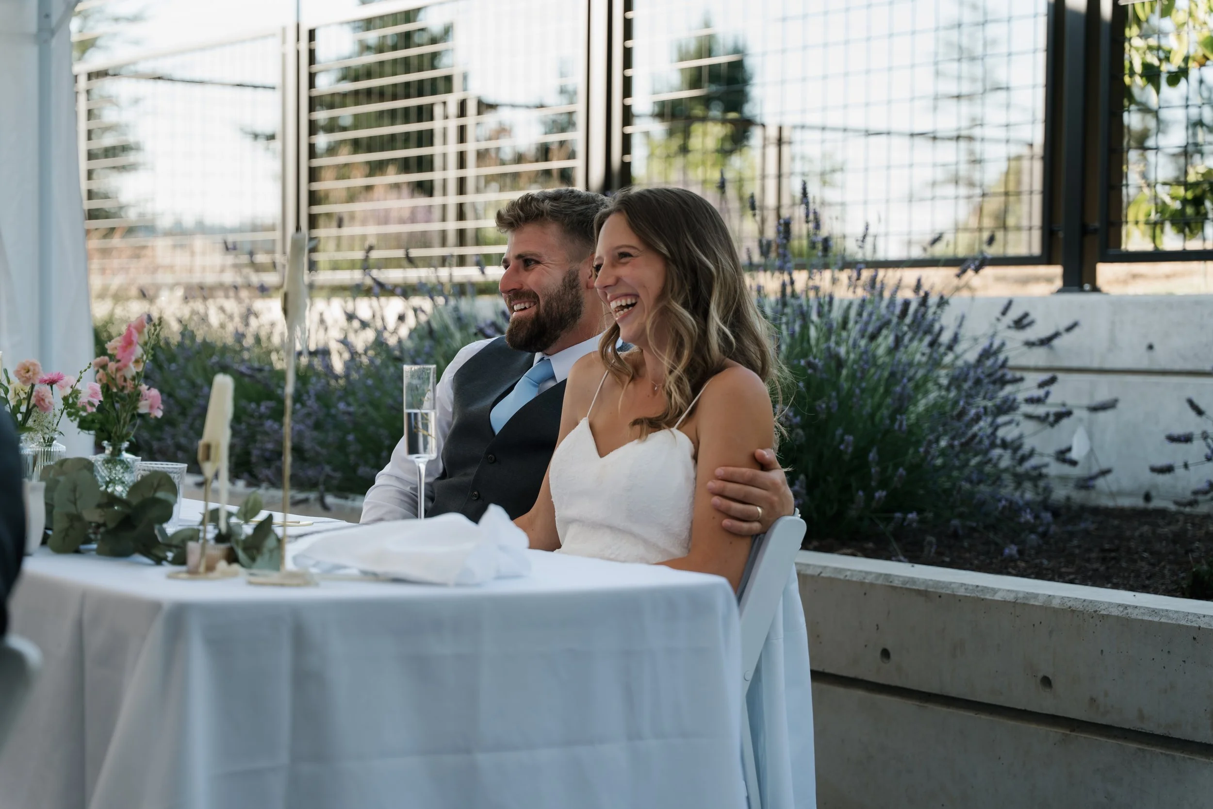 A smiling couple at a wedding reception, sitting at a table with white tablecloth, flowers, candles, and champagne glasses, outdoors with a metal fence and plants in the background.