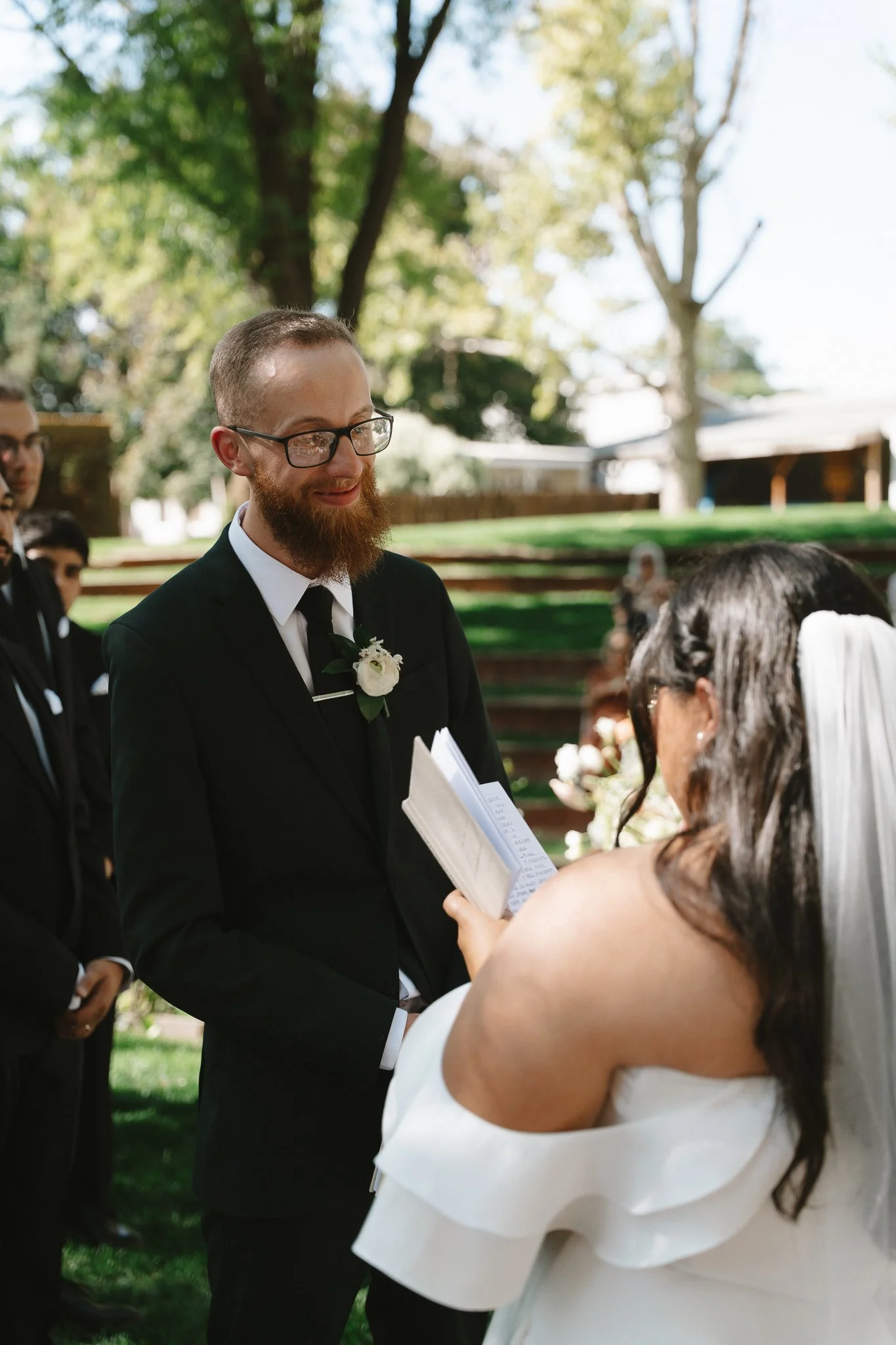 A groom with glasses and a beard wearing a black suit and white boutonniere faces a bride with long dark hair and a veil, who is reading vows from a booklet during an outdoor wedding ceremony in a park with green trees in the background.