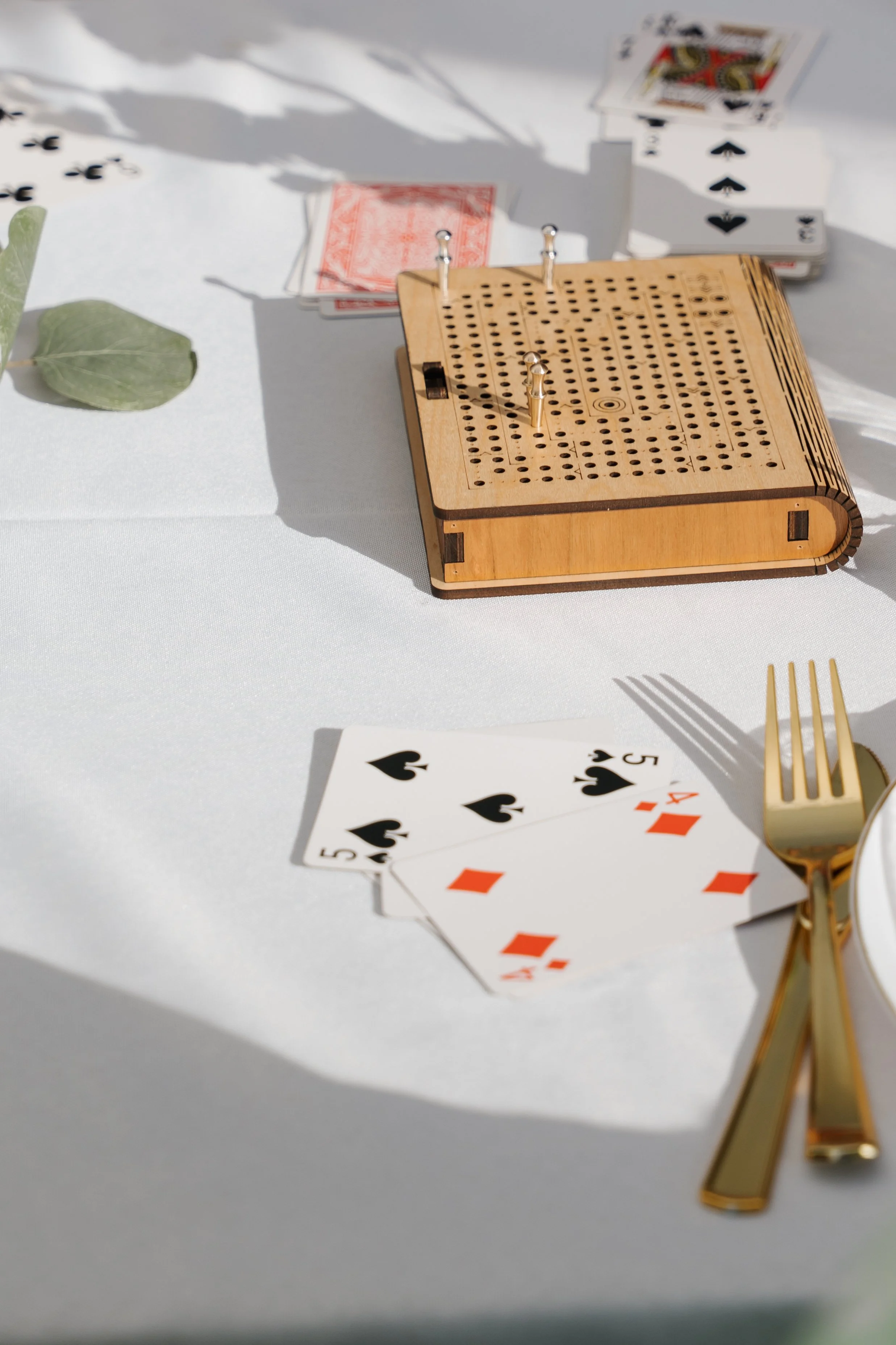 A game of cribbage with playing cards, a wooden cribbage board, and golden fork and knife on a white table.