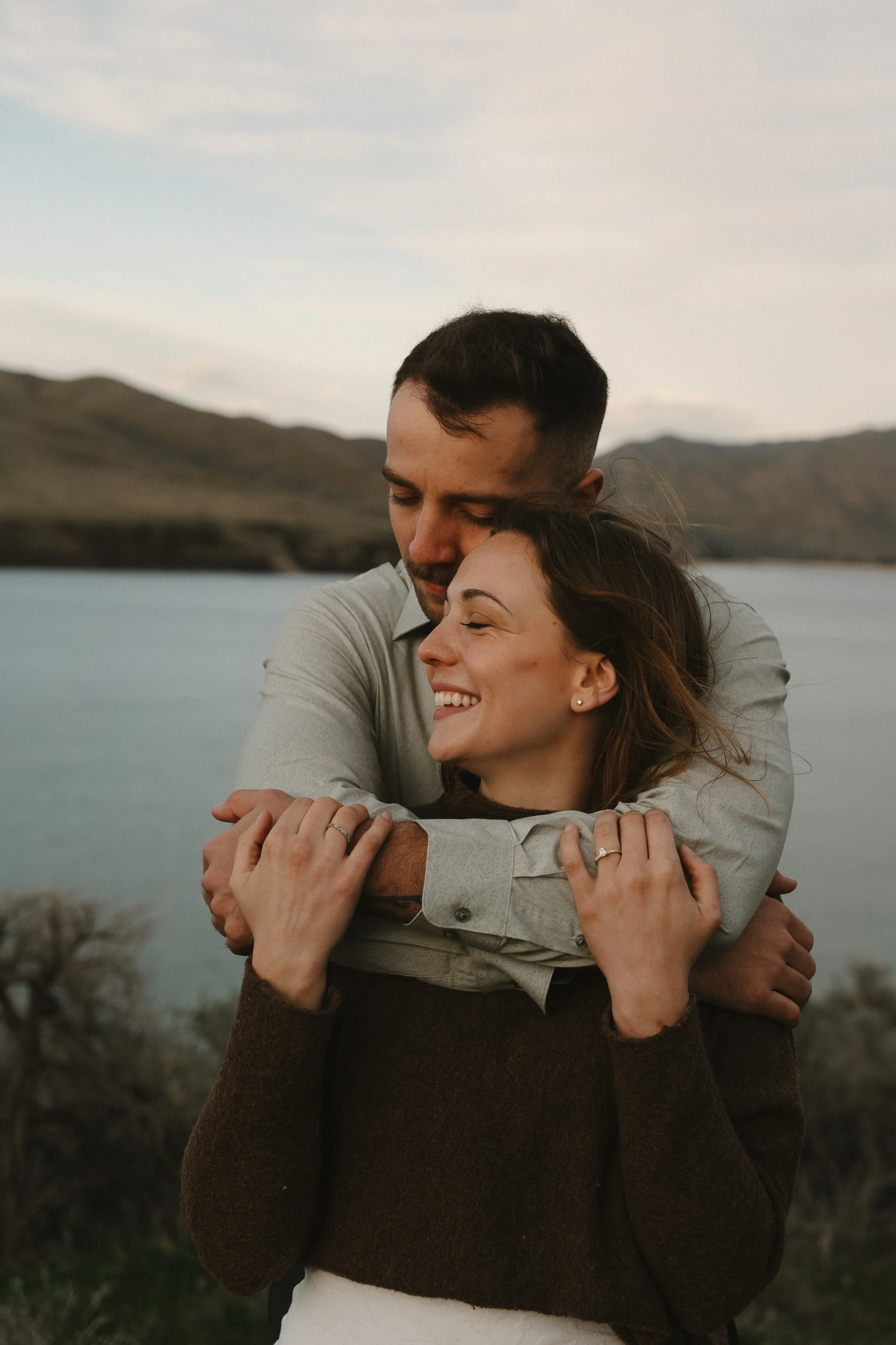 Smiling couple embracing outdoors near a lake with mountains in the background at sunset.