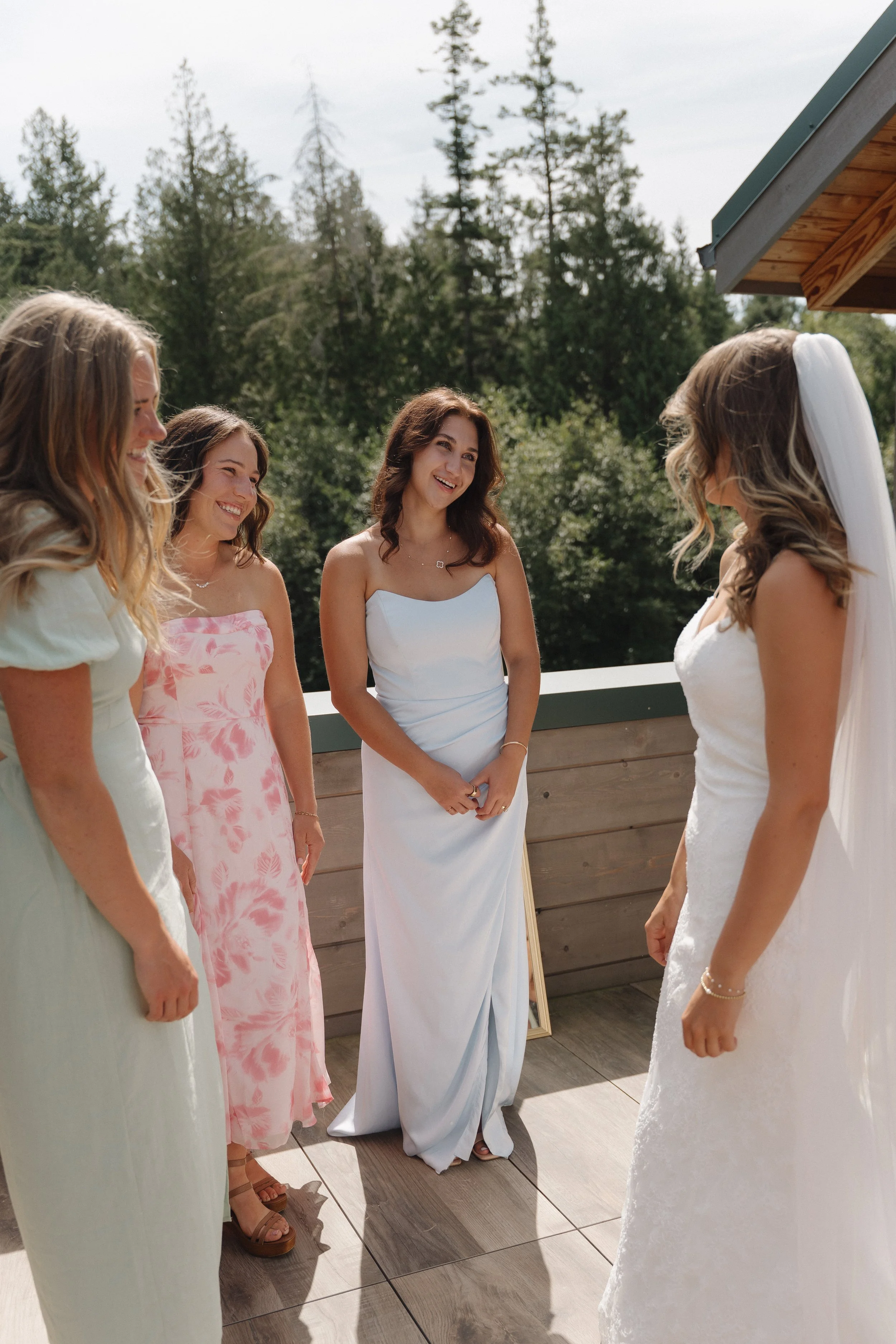 Bride and bridesmaids standing outdoors on a wooden deck, smiling and talking, surrounded by trees, during a wedding celebration.