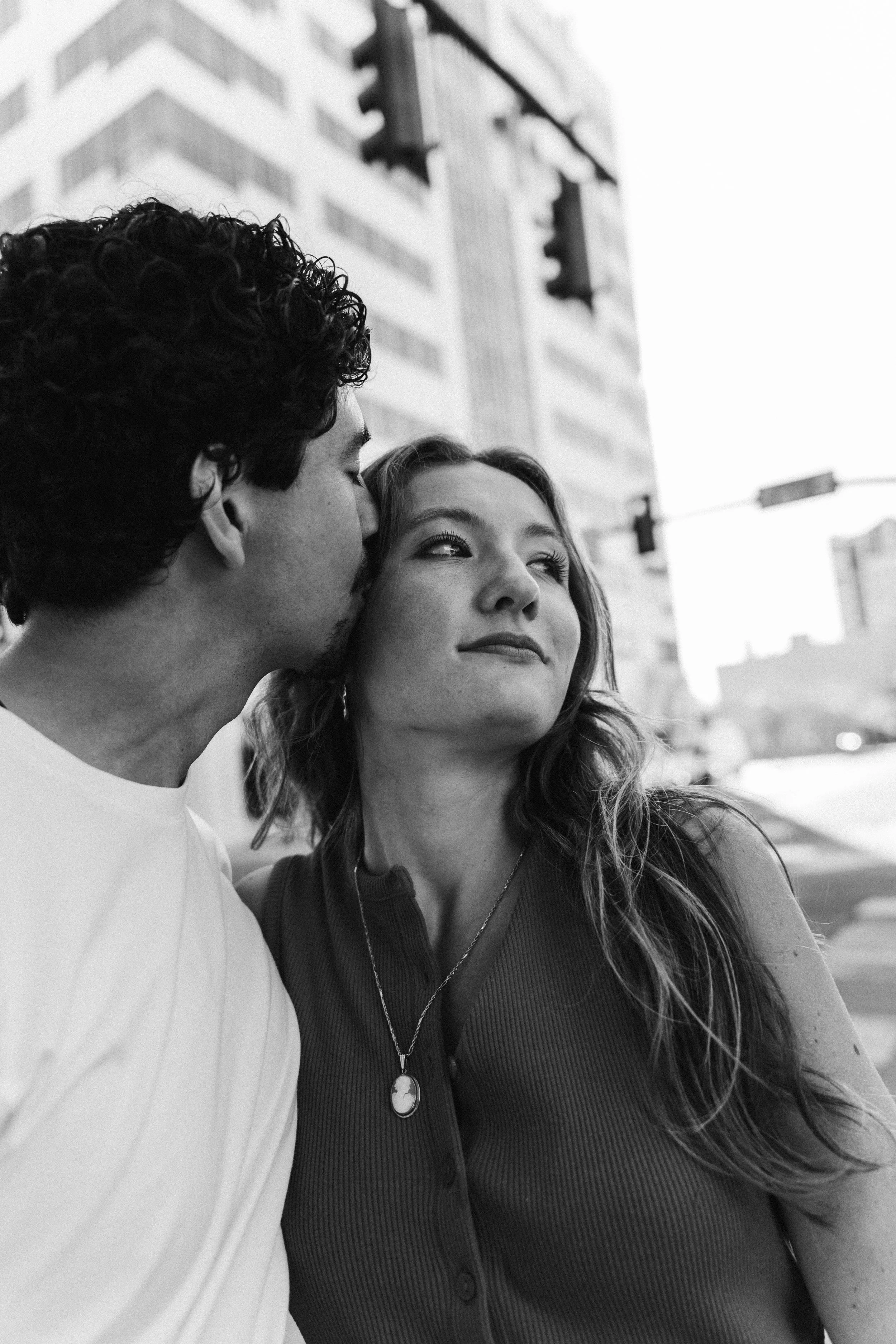A black-and-white photo of a young man kissing a woman on the temple on a city street, with tall buildings and traffic lights in the background.