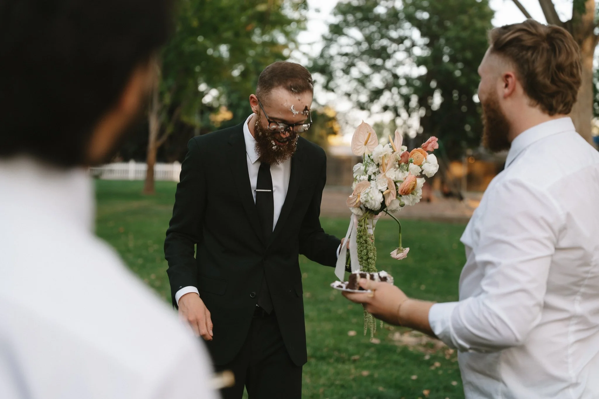 A man in a black suit with a beard and glasses is smiling and covered in cake, standing outdoors with trees in the background. Another man with a beard and a white shirt is holding a plate with a slice of cake and a bouquet of flowers, celebrating a 