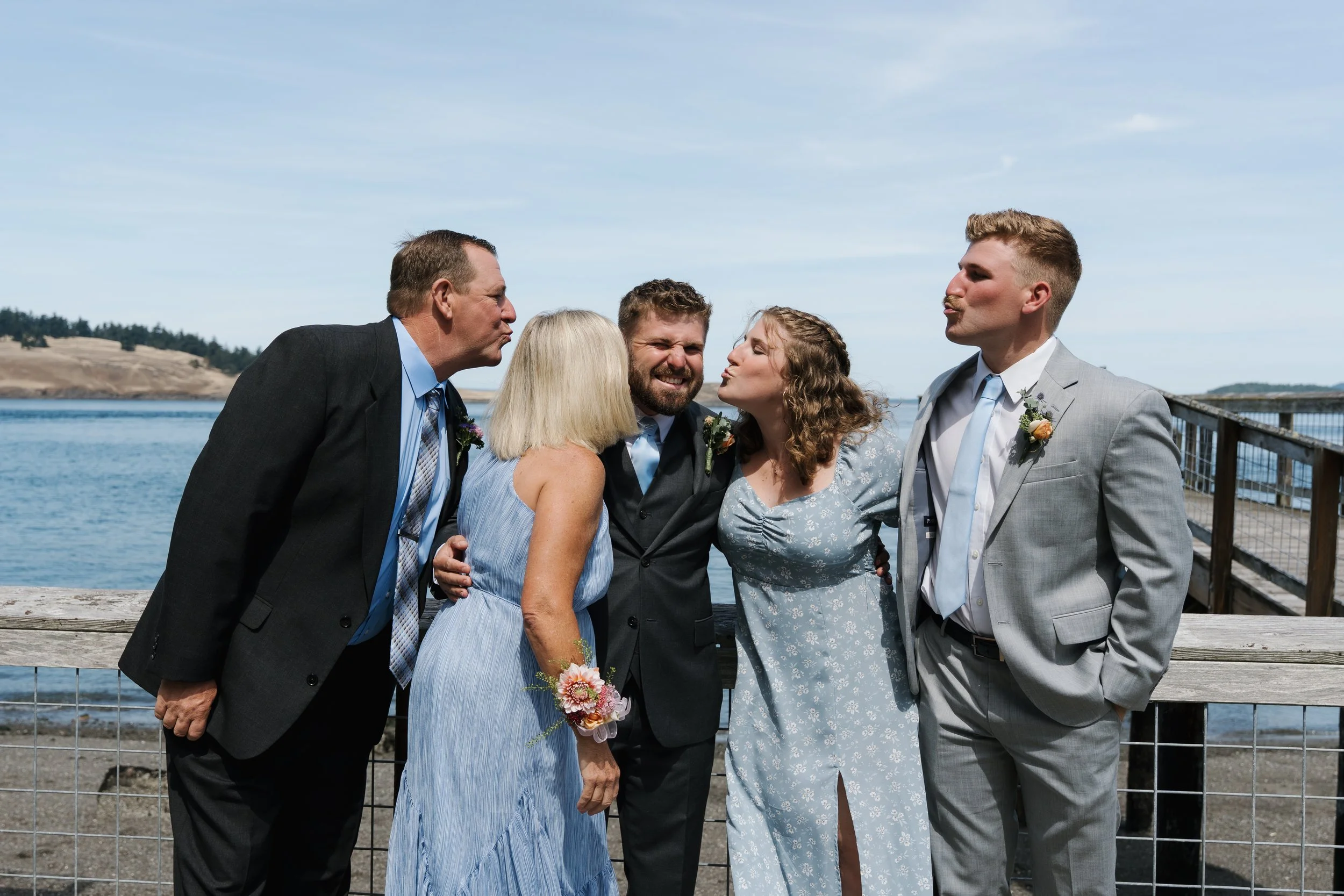 A group of five people posing for a photo on a pier near the water, dressed in formal attire. The two women are in dresses, and the men are in suits. They are making puckered lips while leaning toward each other, with a scenic ocean and hilly landsca