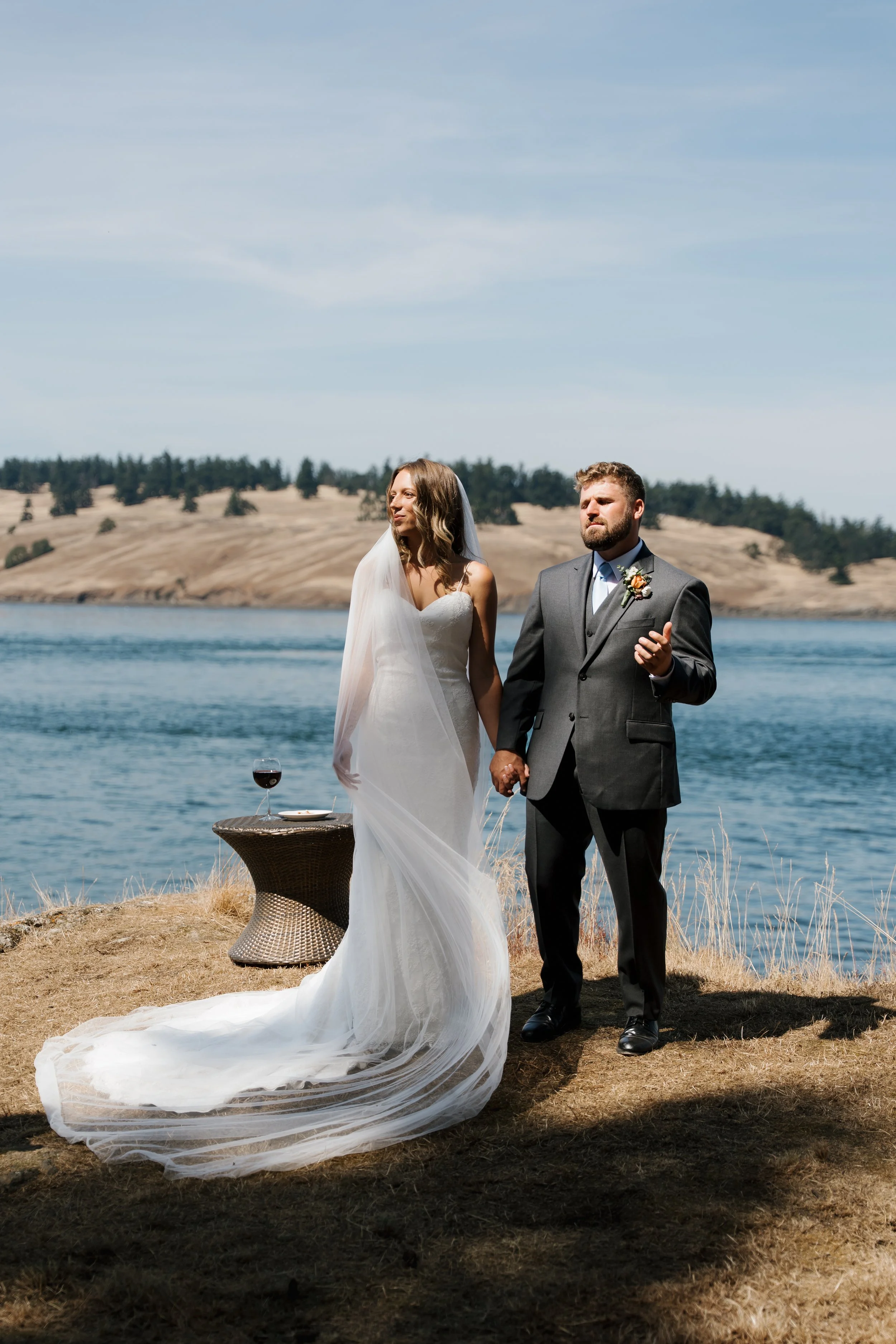 Bride and groom exchanging vows outdoors by a lake with a scenic background of hills and trees, holding hands, with a glass of red wine and a plate on a small table beside them.