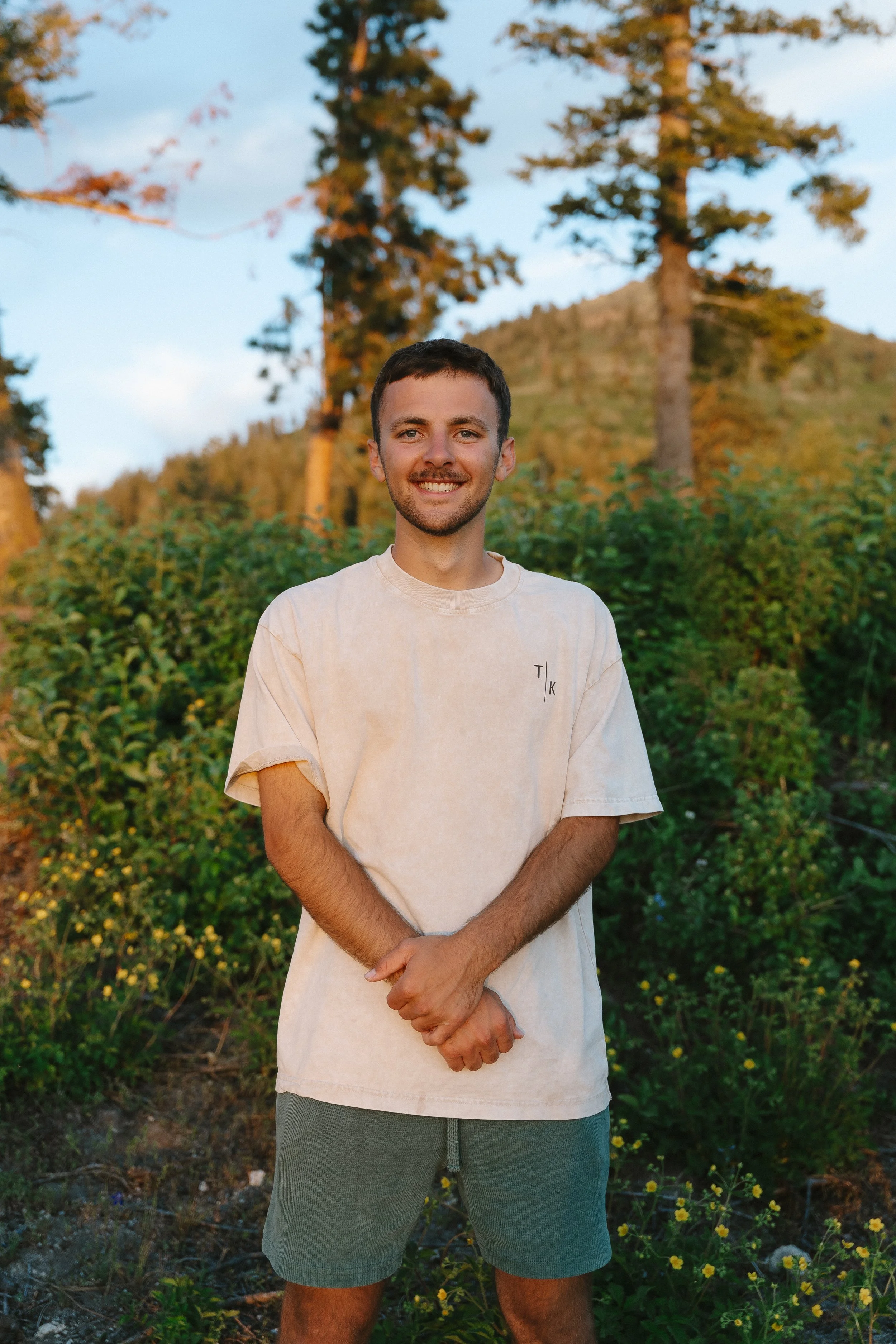 A young man with short brown hair and a beard smiling outdoors, standing in front of green bushes and trees with mountains and a blue sky in the background.