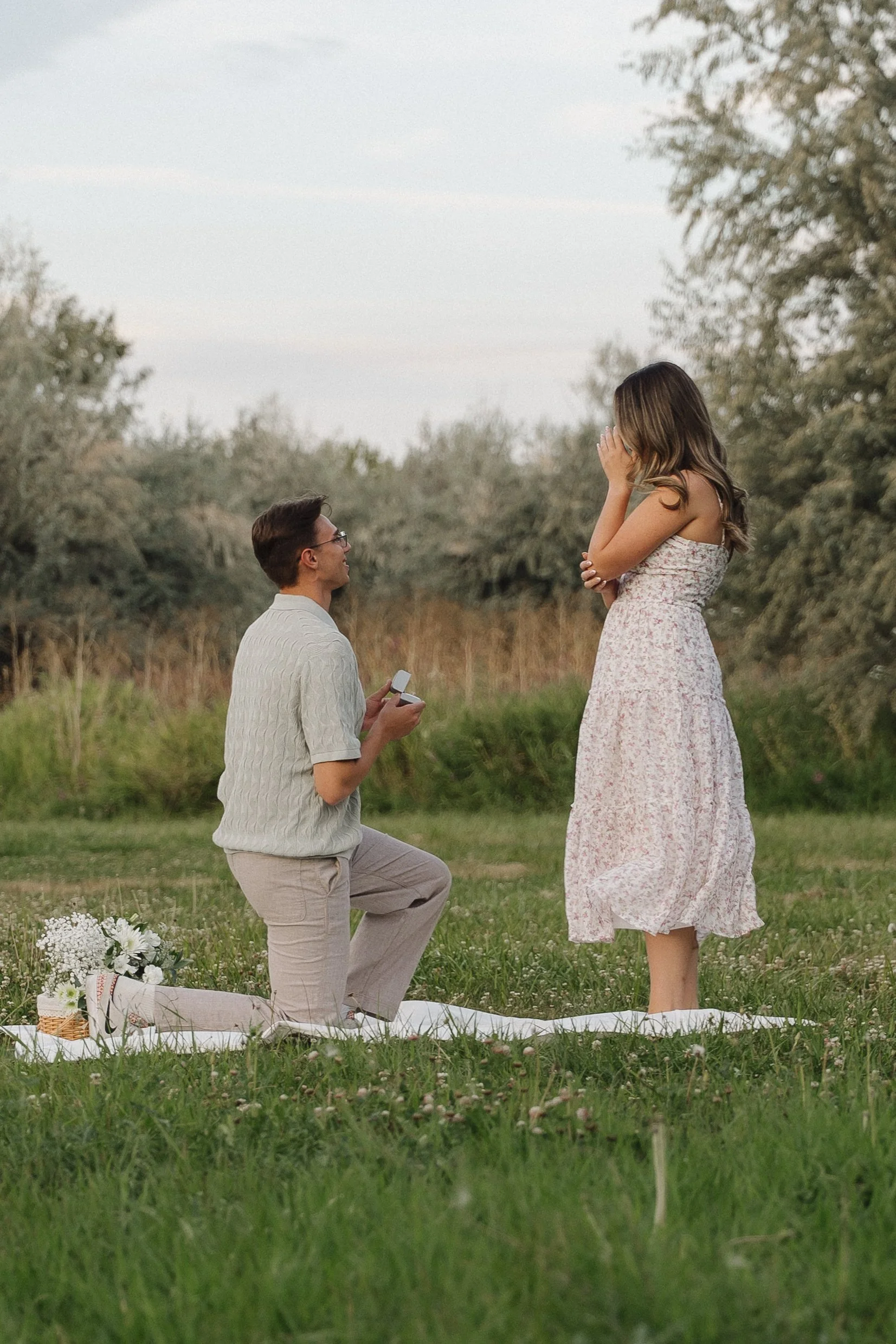 A man kneeling on one knee proposes to a woman in a grassy outdoor setting during daytime, with a bouquet of flowers nearby.