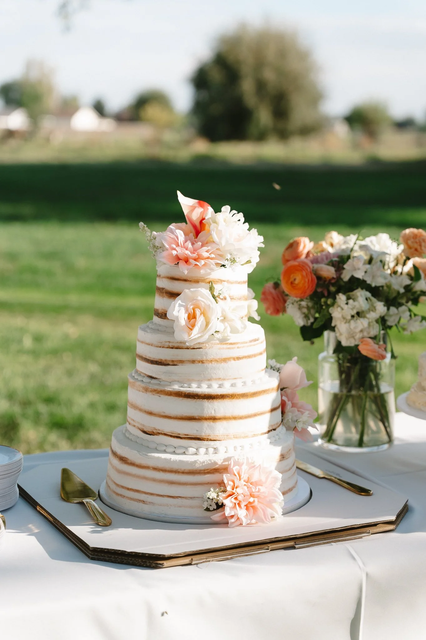 A three-tiered naked wedding cake decorated with peach and white flowers, placed on a white table with gold utensils, beside a floral arrangement in a glass vase, outdoors on a sunny day.