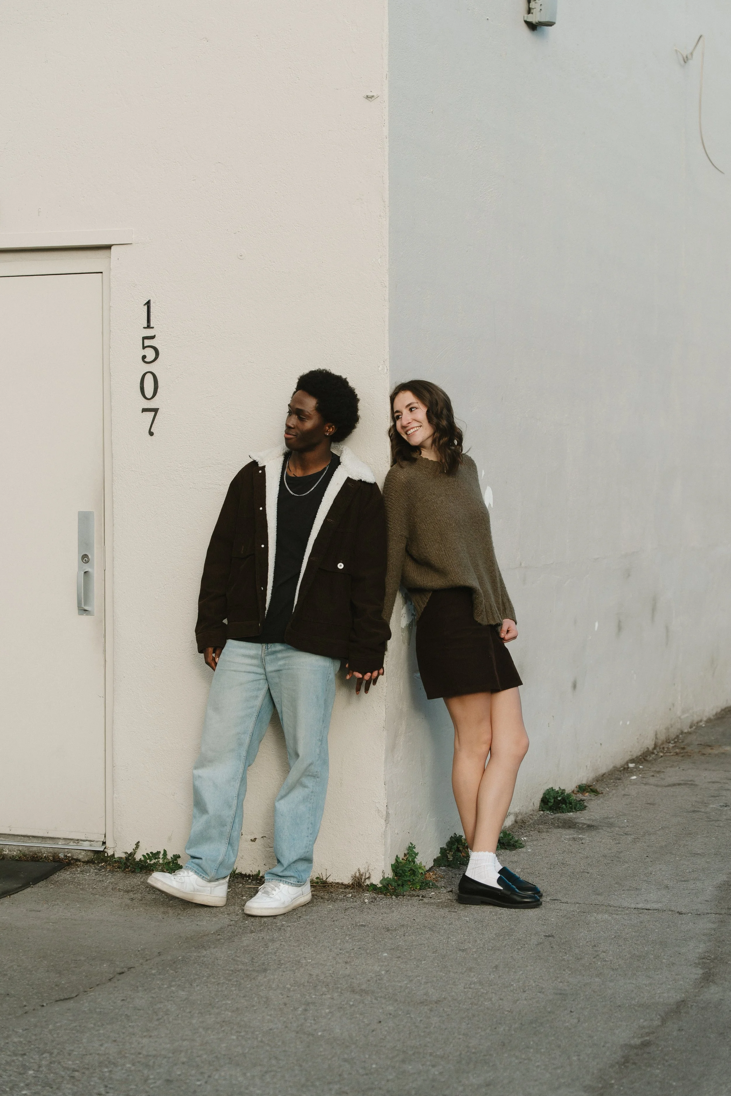 A young man and woman casually leaning against a corner of a white and grey wall, holding hands and smiling.