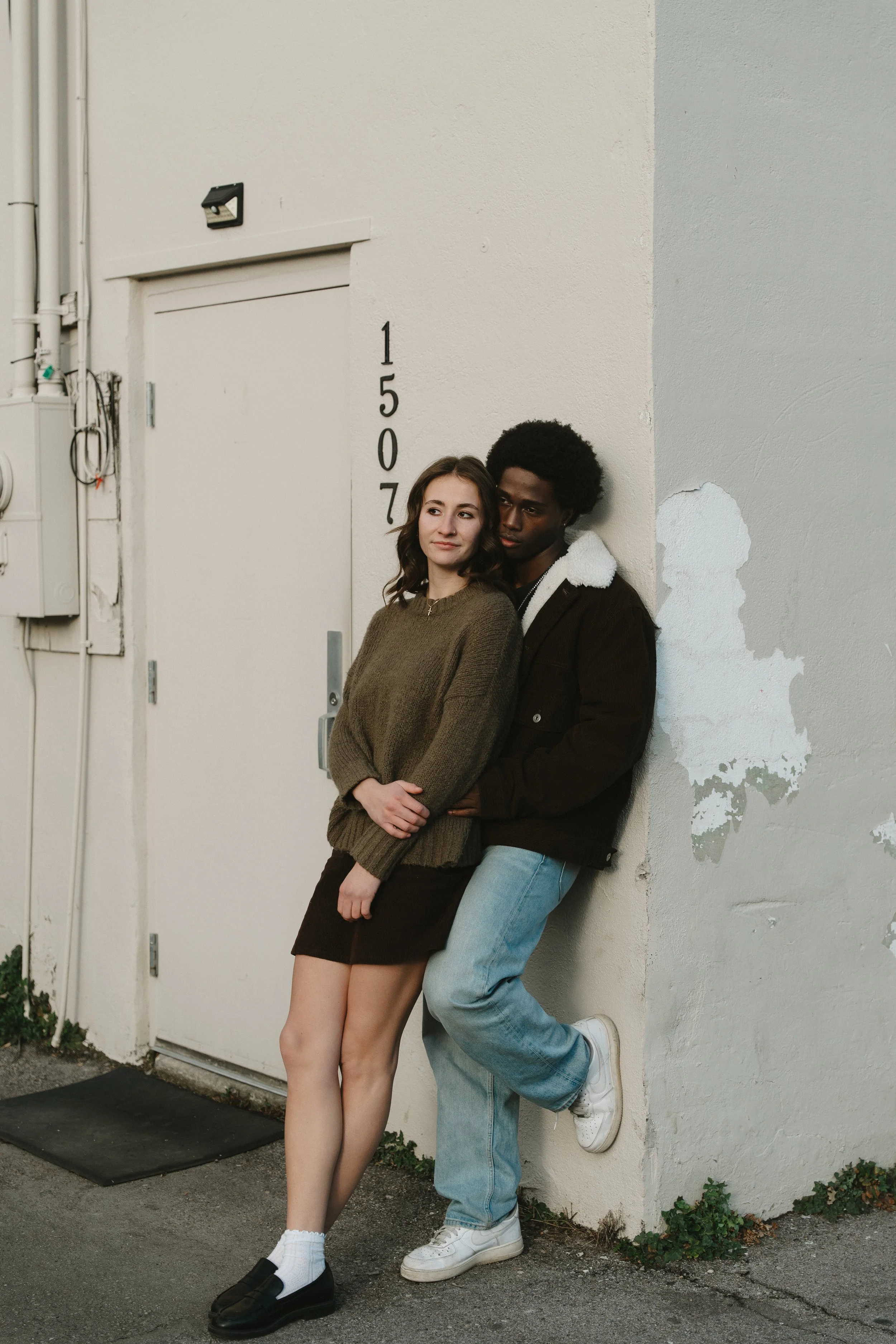 A young woman and man leaning against a wall outside a building numbered 1507, with the woman in a brown sweater and skirt and the man in a brown jacket and jeans.