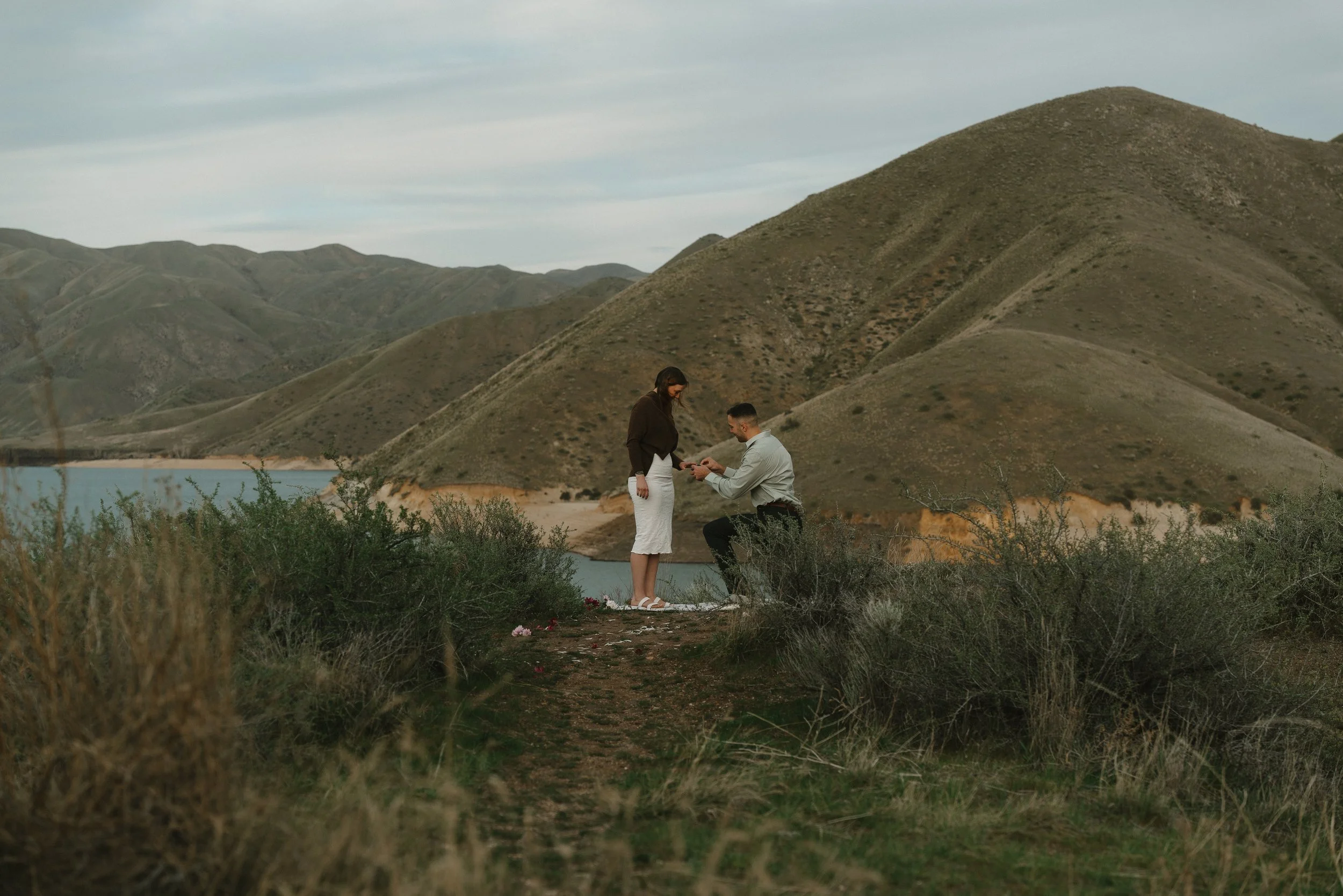 A man proposing marriage to a woman in a scenic outdoor location with mountains and a body of water in the background.