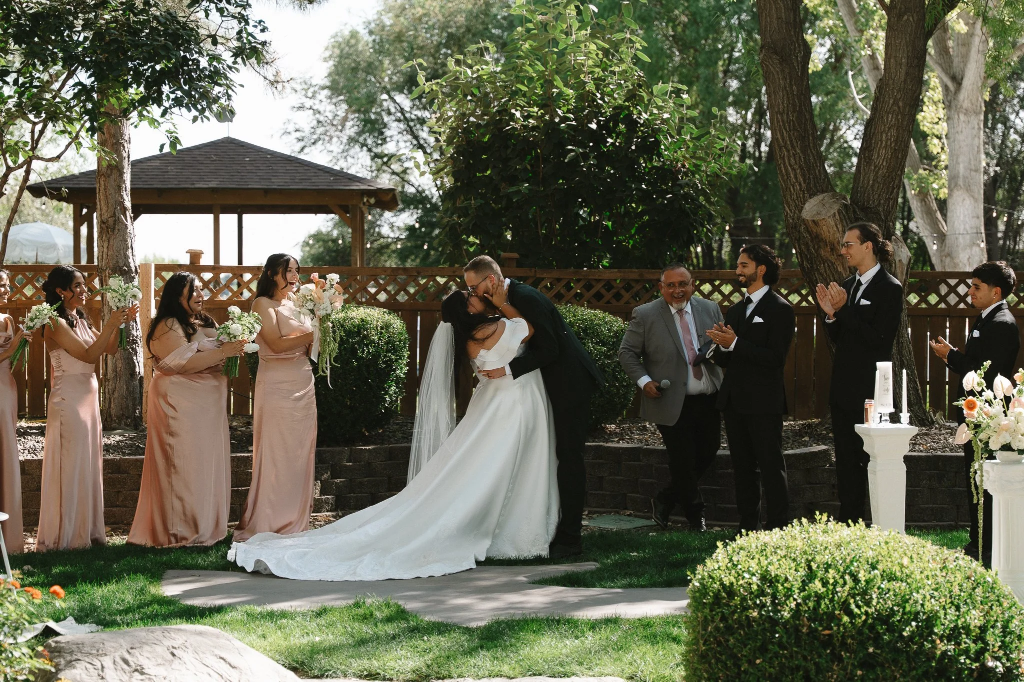A couple getting married outdoors, with the bride and groom kissing, surrounded by bridesmaids and groomsmen, and a person holding a microphone smiling nearby.