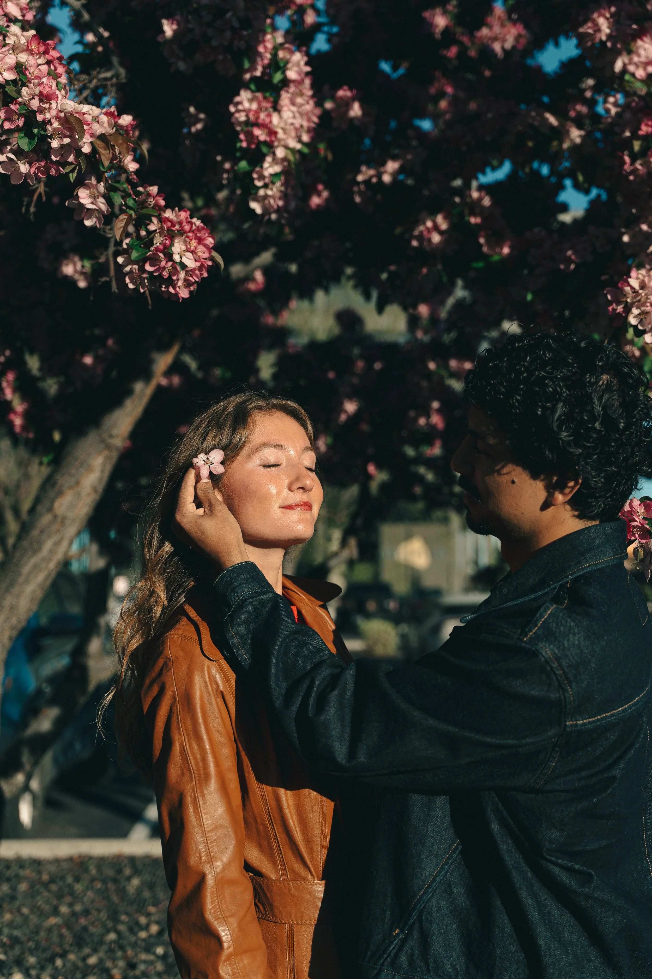 A man gently touches a woman's face with a flower in her hair in front of a blooming pink tree.