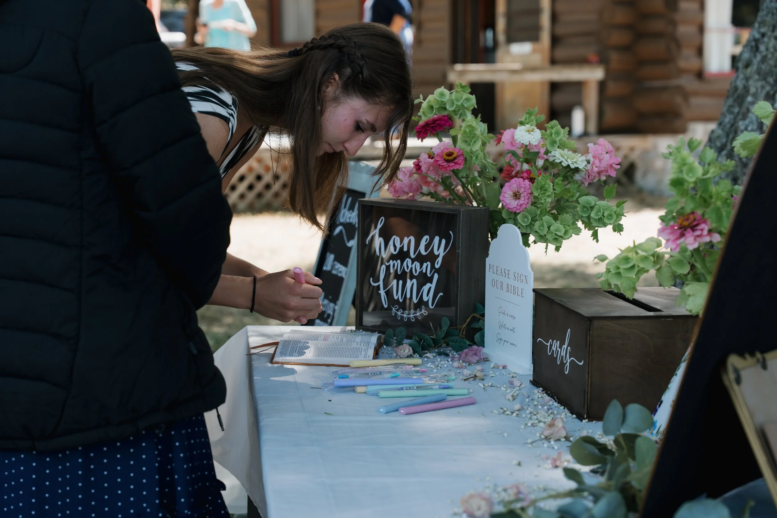 A young woman writes on a card at a wedding or event sign-in table decorated with flowers, signs, and pens.