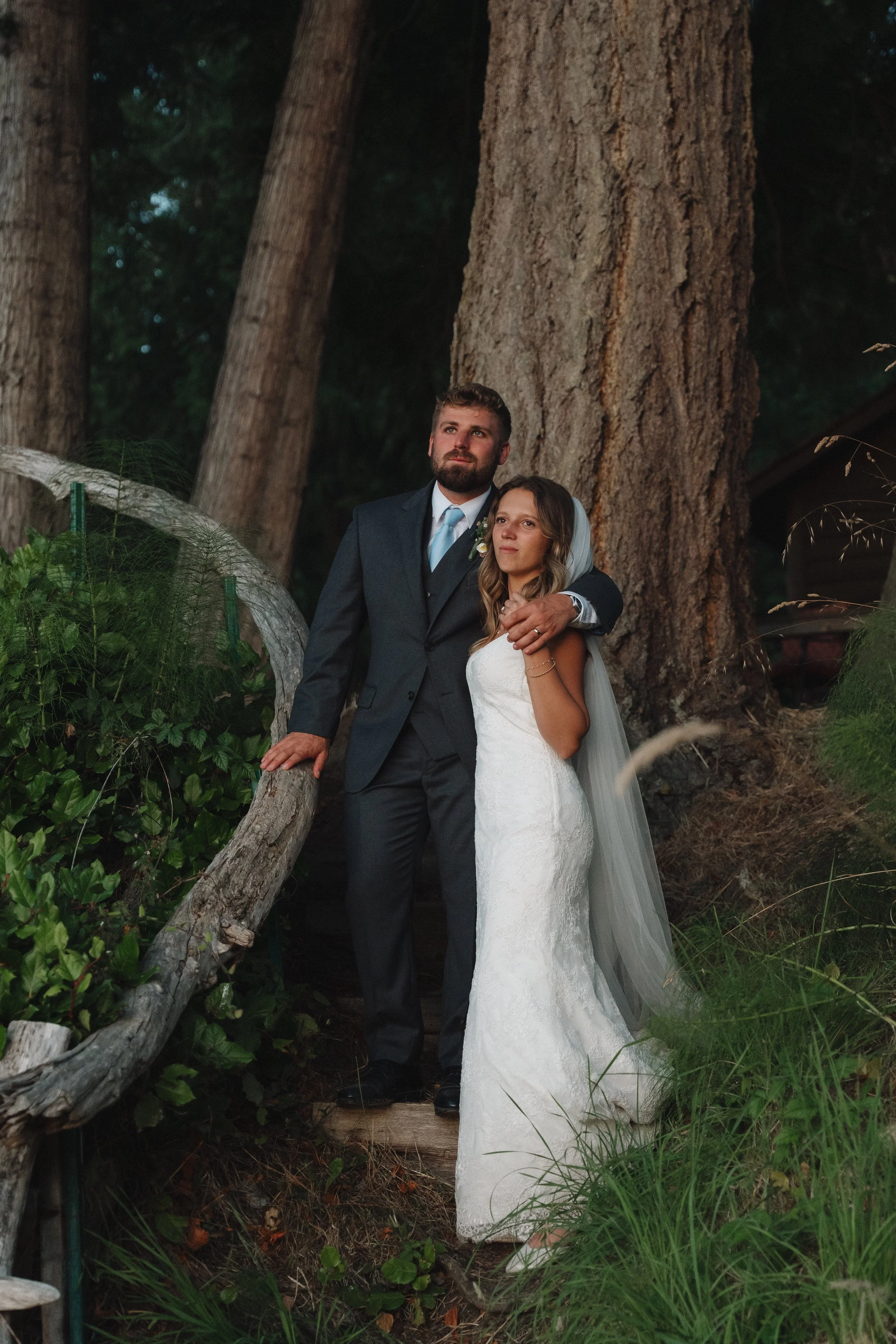 A bride and groom standing outdoors in front of tall trees, with the groom's arm around the bride's shoulders and both looking into the distance.