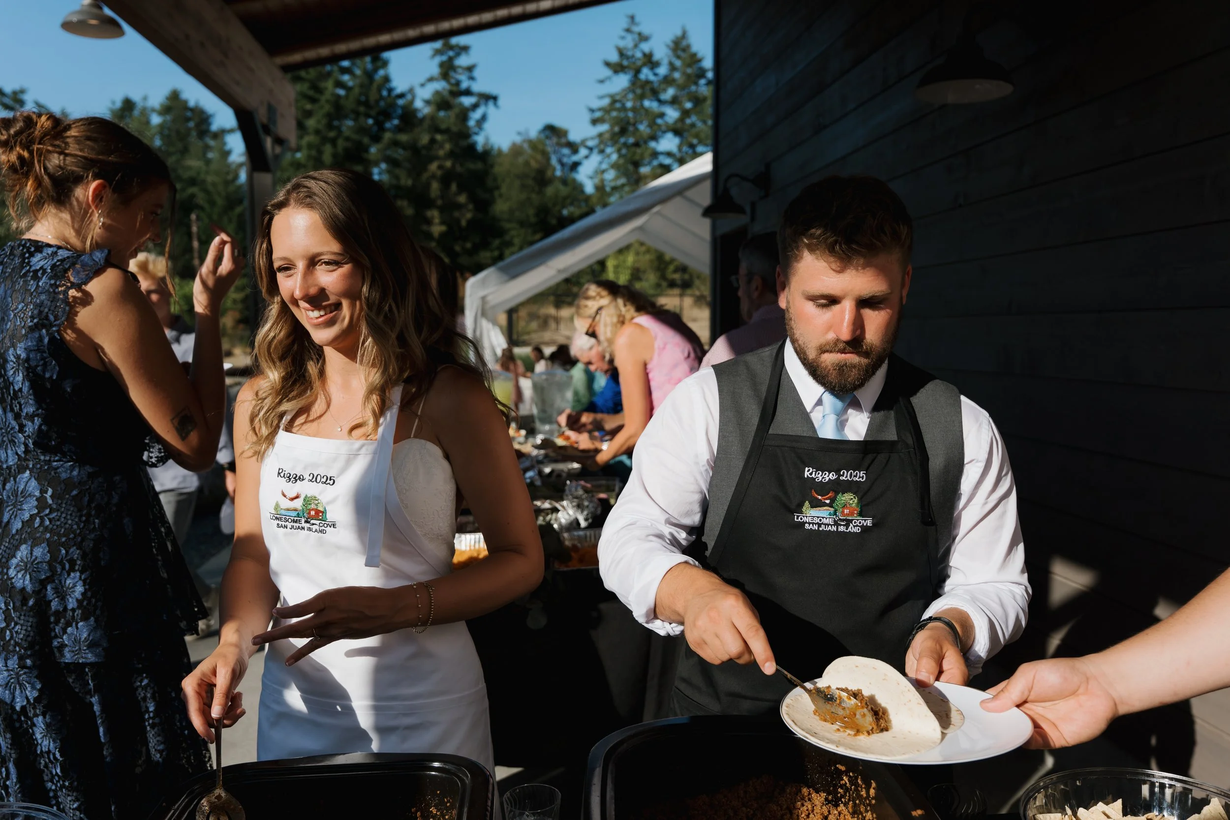 People serving and enjoying food at an outdoor gathering, with a woman in a white dress smiling and a man in a white shirt and black apron serving tacos, in a sunny outdoor setting.