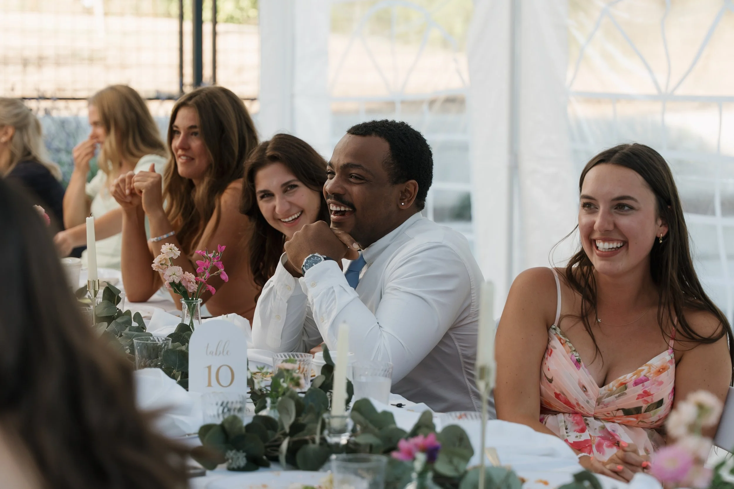 People enjoying a wedding reception, sitting at a decorated table with flowers and candles, smiling and laughing.