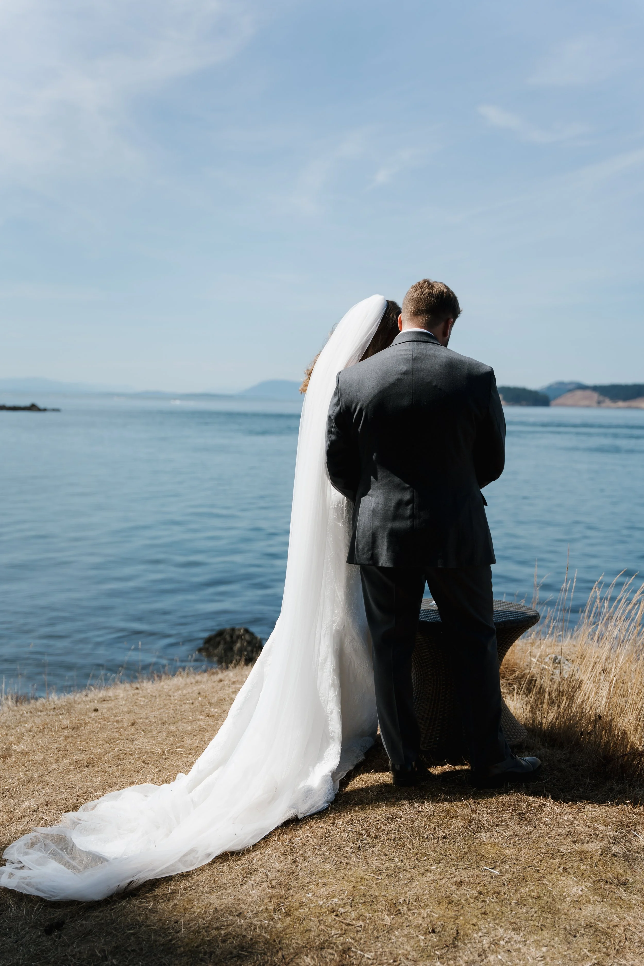 A bride in a white wedding dress and veil standing next to a groom in a suit near the water, with mountains in the distance under a blue sky.