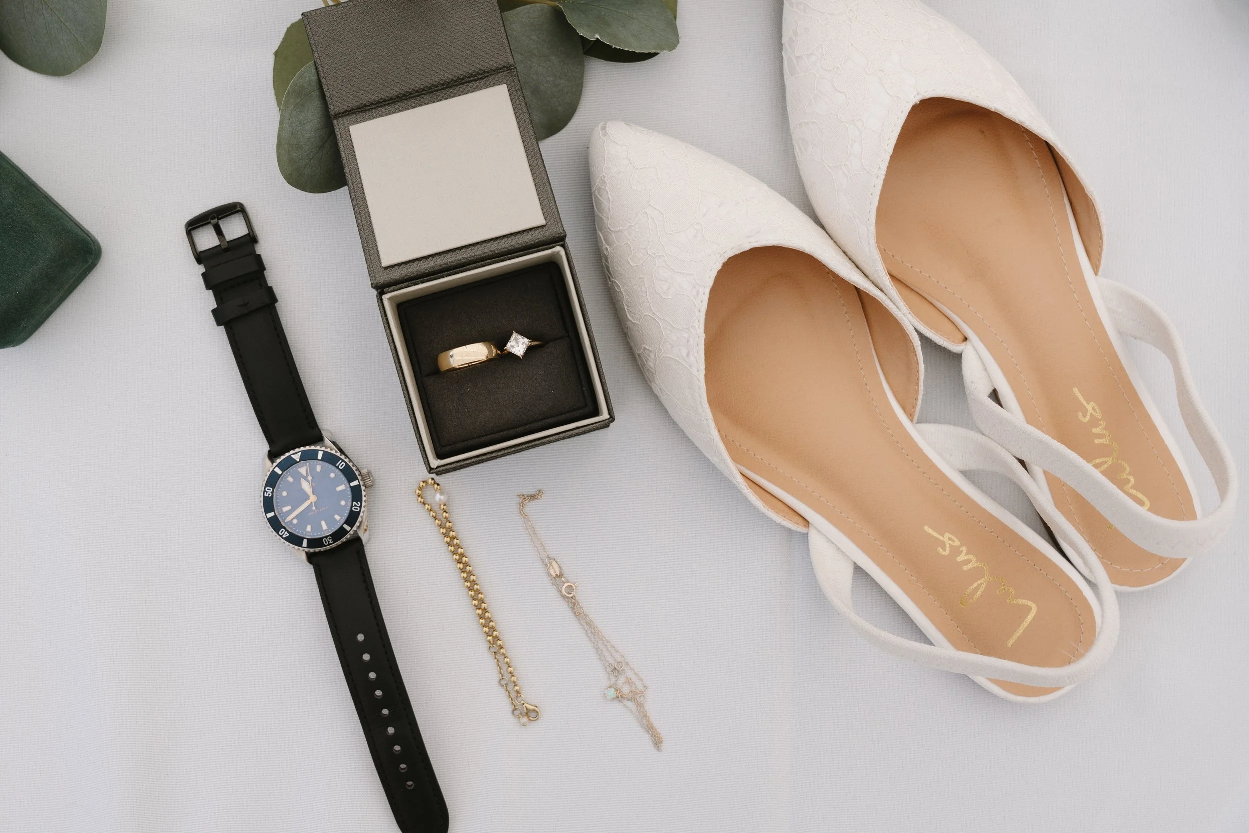 A collection of wedding accessories, including a pair of white lace high heel shoes, a black wristwatch, a gold bracelet, a gold and silver necklace, a ring in a black jewelry box, and a black watch strap, all arranged on a white surface.