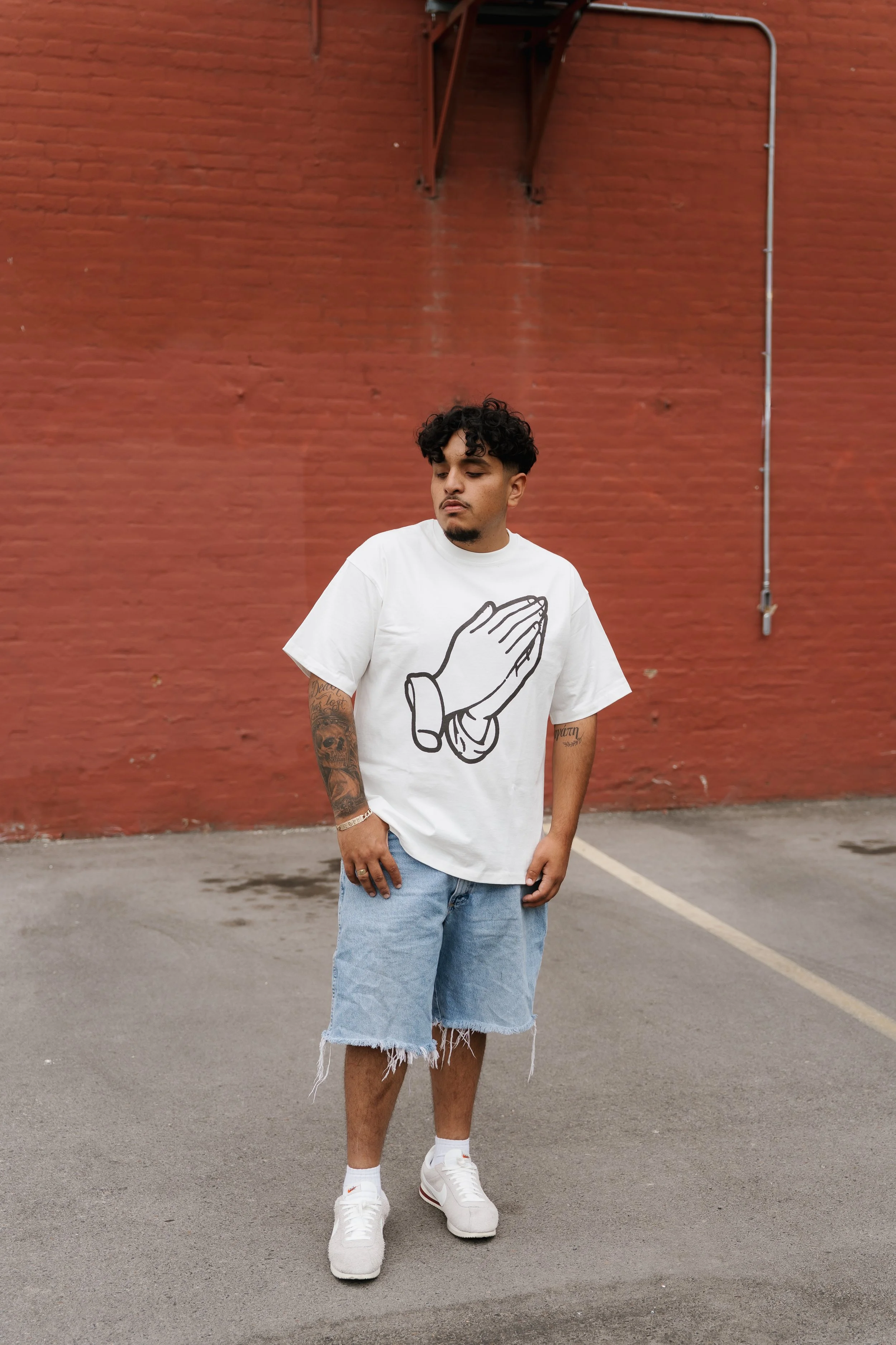 A young man standing in front of a red brick wall, wearing a white T-shirt with a large praying hands graphic, light denim shorts, white sneakers, and sporting tattoos on his arms.