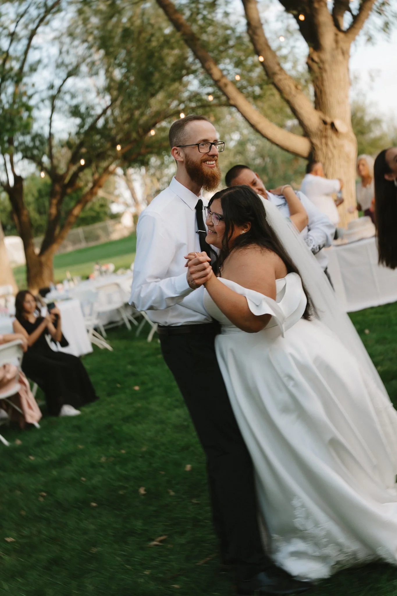 Couple dancing at an outdoor wedding reception, with guests seated at decorated tables and string lights hanging from trees in the background.