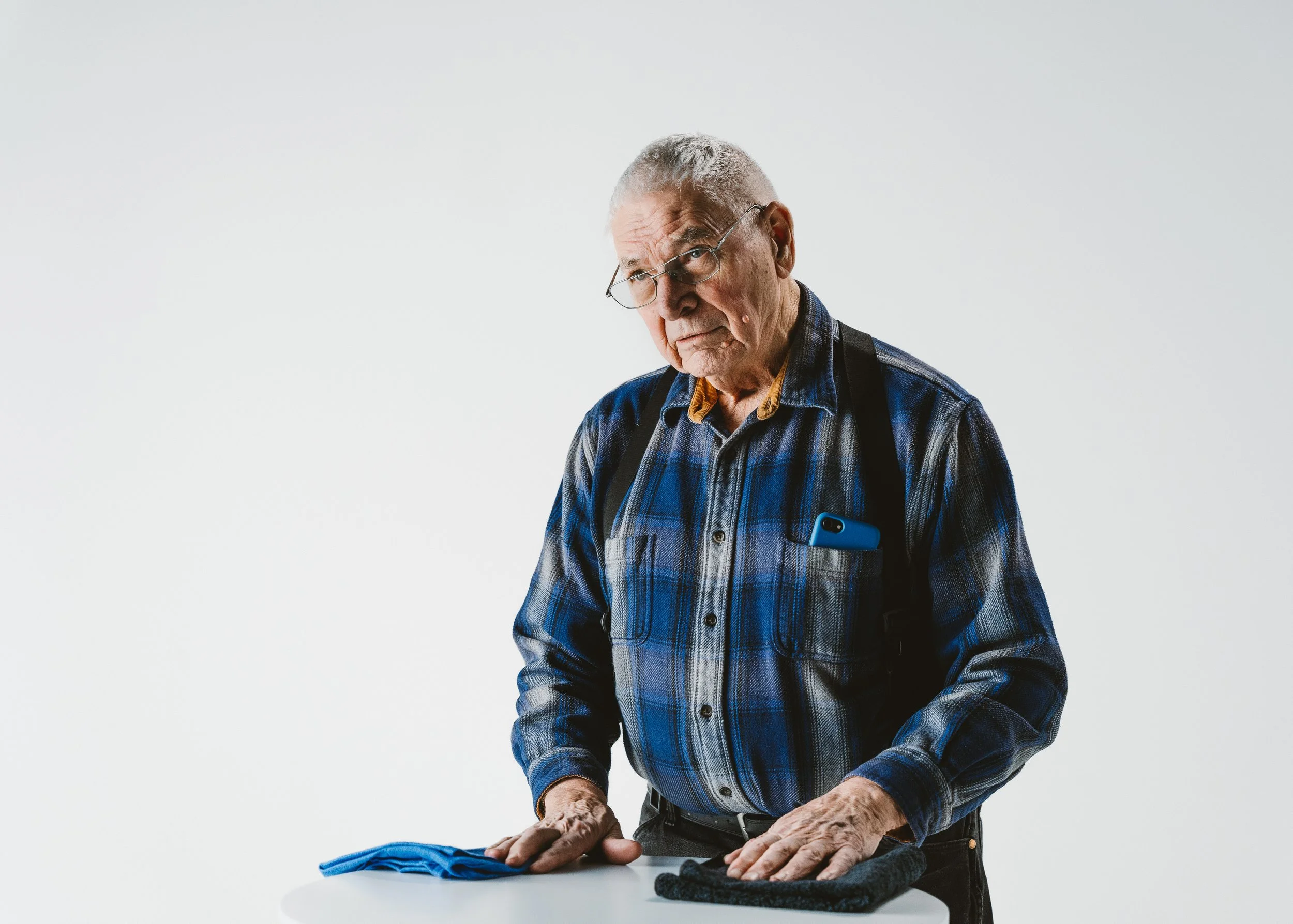 An elderly man with glasses and gray hair, wearing a plaid shirt with a phone in his chest pocket, stands at a white table with a blue towel and black gloves, looking confused or concerned.  He is being photographed for a commercial 
