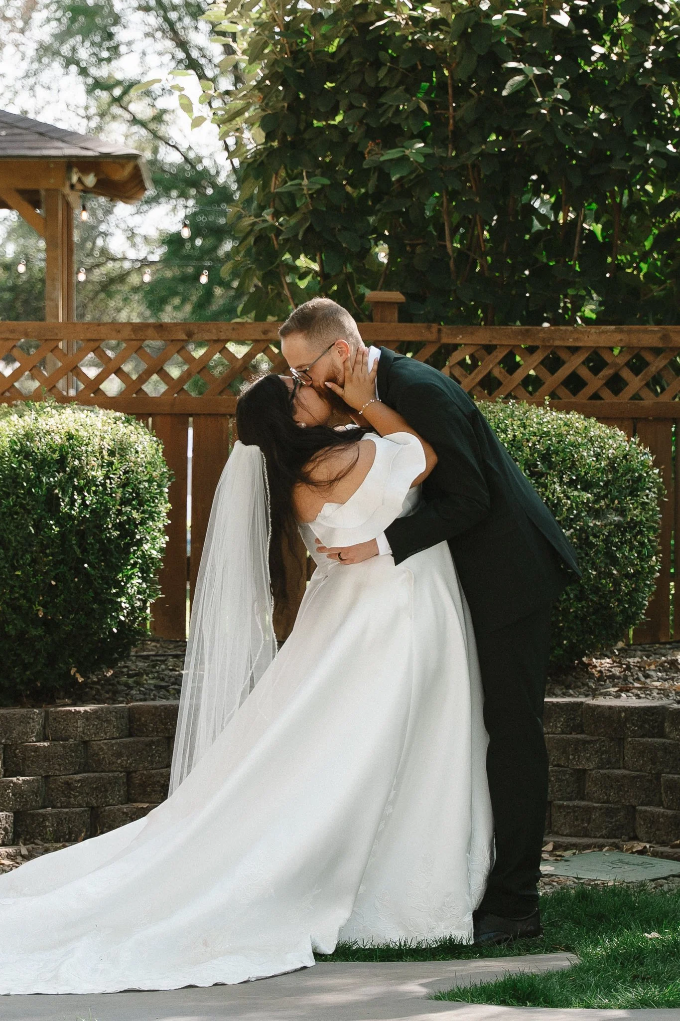 A bride and groom sharing a kiss outdoors, with the groom bending slightly and holding the bride's waist, in front of a wooden fence and green bushes.