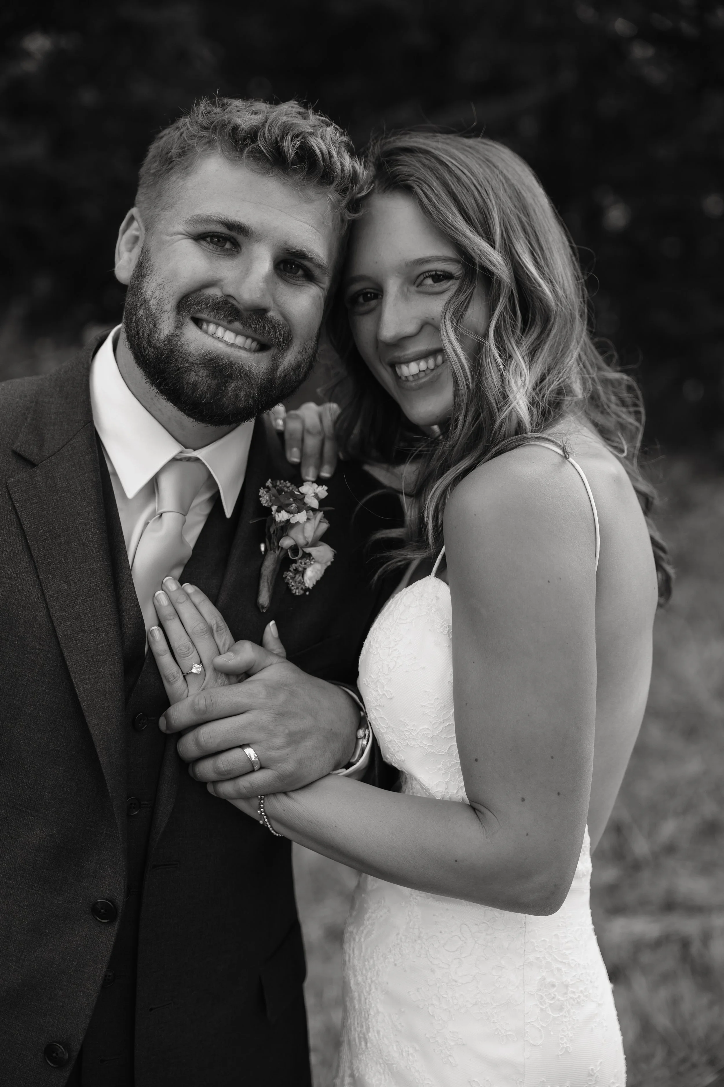 Black and white photo of a wedding couple smiling, with the bride wearing a strapless wedding dress and the groom in a suit with a boutonniere, standing outdoors.