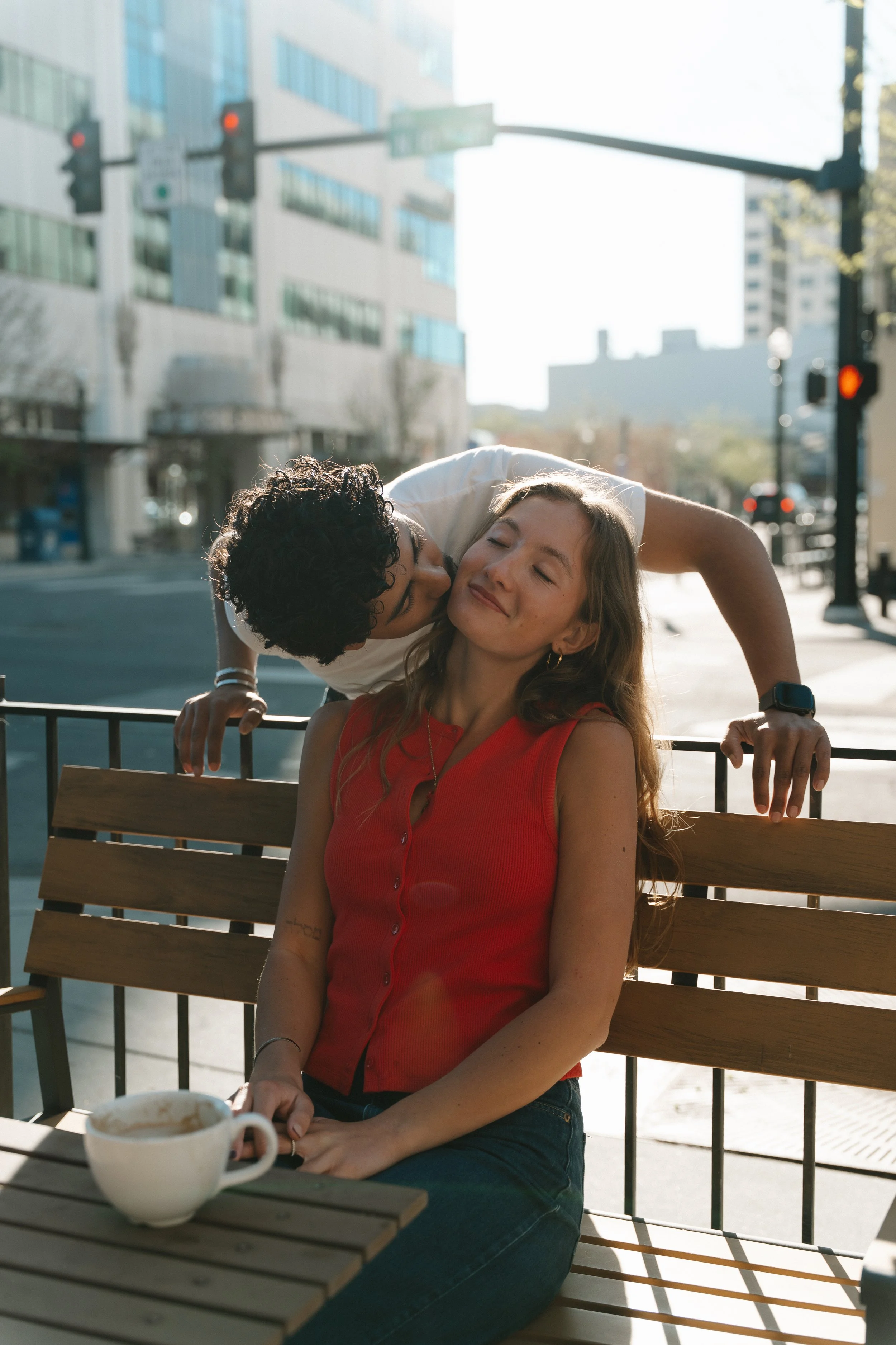 A woman in a red sleeveless top sitting on a park bench with a coffee cup in front of her, smiling with eyes closed as a man behind her gives her a kiss on the cheek in an urban setting.