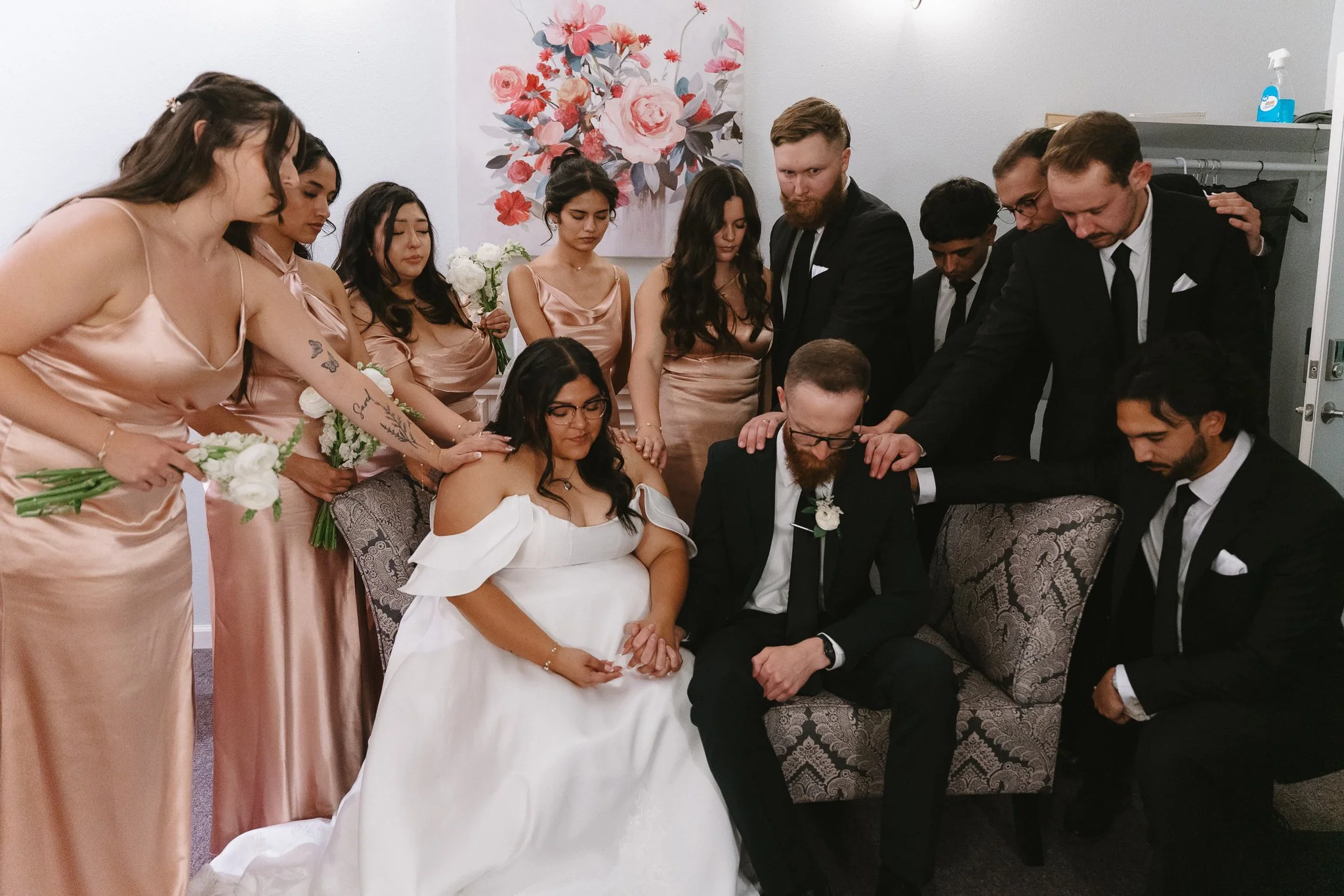 A group of people participating in a wedding or spiritual ceremony, with the bride and groom seated, surrounded by bridesmaids and groomsmen in a room decorated with a floral painting.