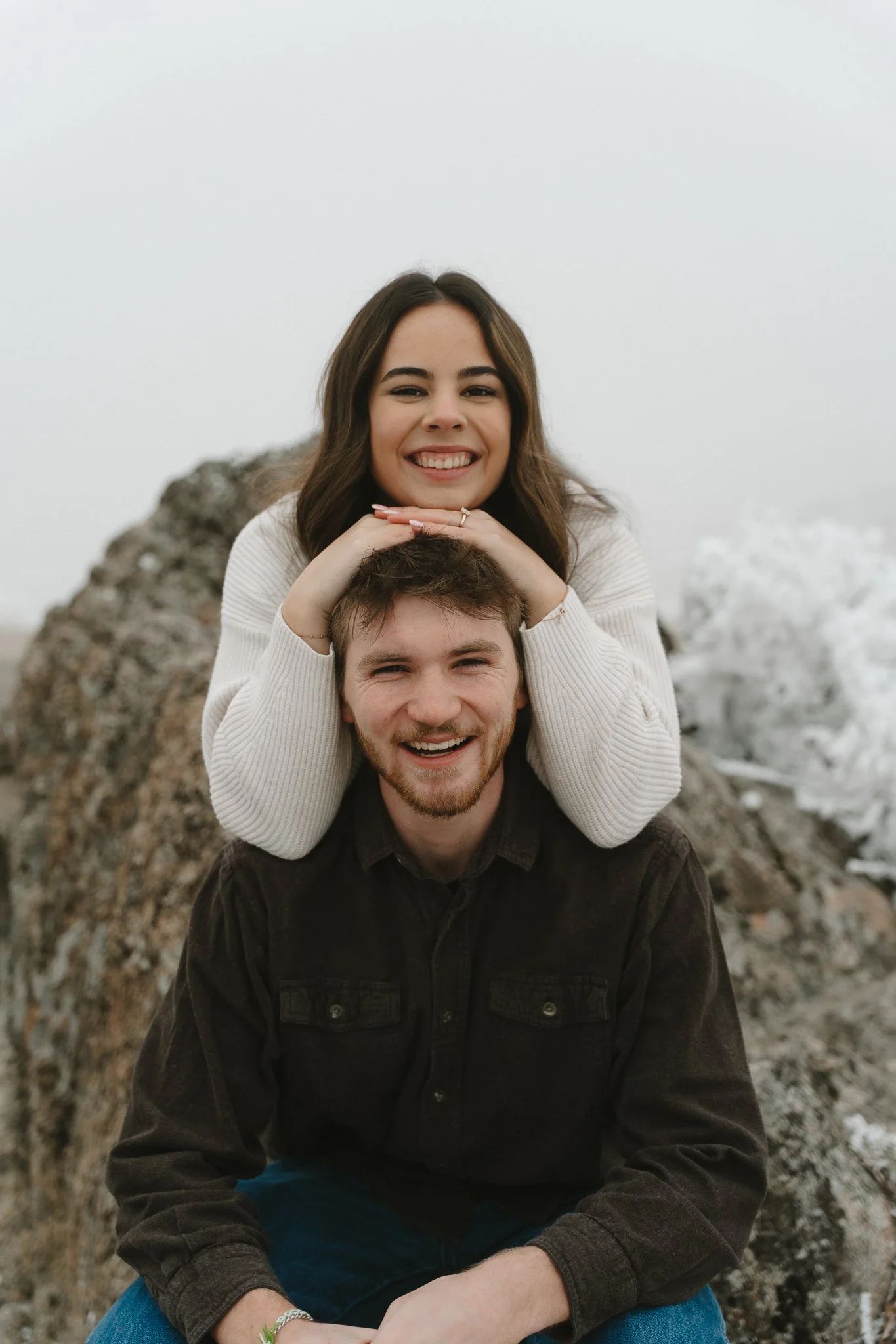 A happy young couple outdoors, with one woman sitting on the man's shoulders, both smiling, near rocks with snow in the background.