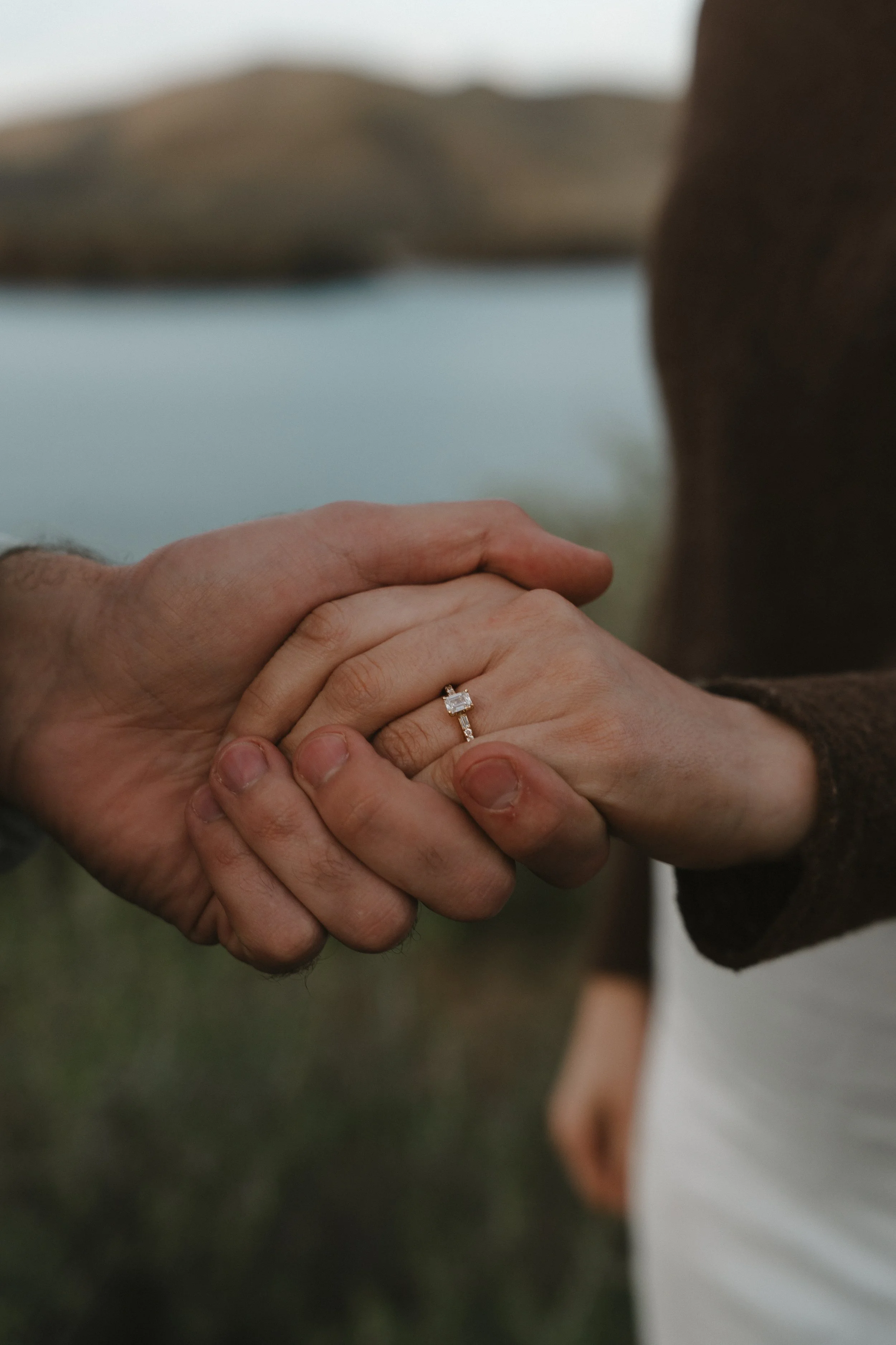 Close-up of a couple holding hands, with a focus on a woman's hand wearing an engagement ring, in an outdoor setting near a body of water and hills in the background.