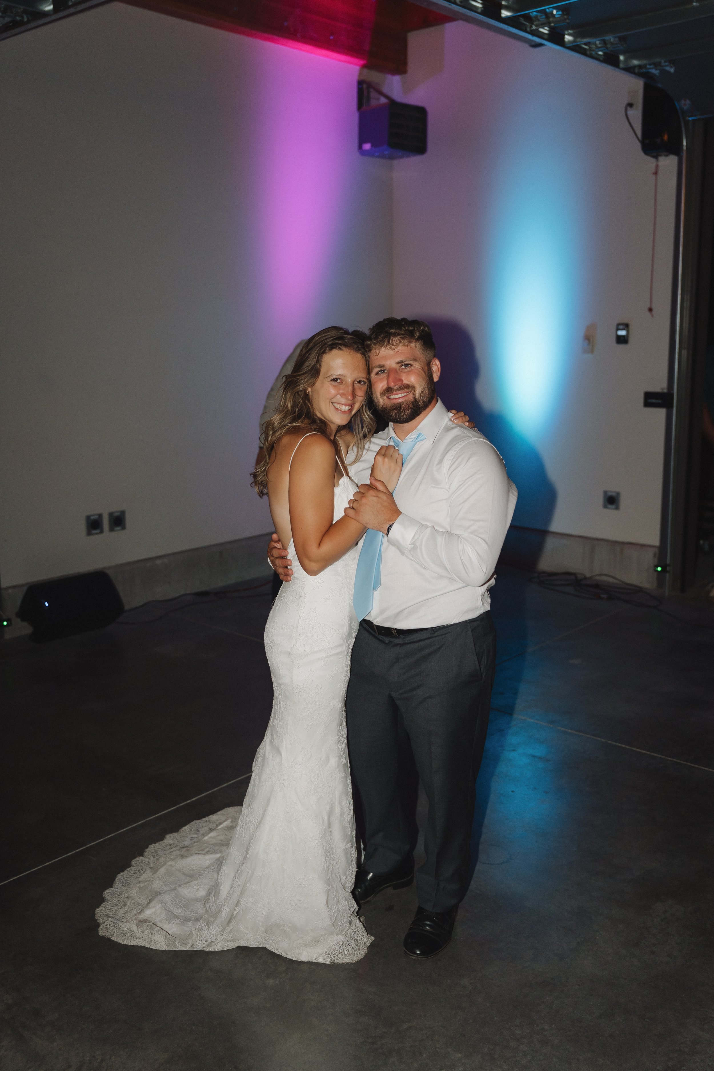 A bride and groom are dancing and smiling during their wedding reception in a dimly lit room with colorful uplighting.