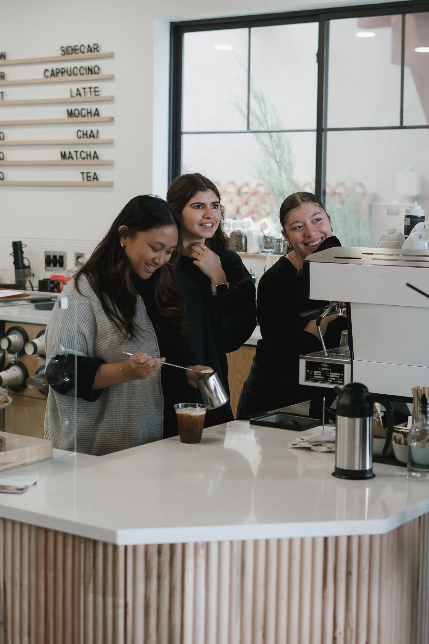 Three women standing at a coffee shop counter, smiling and preparing drinks near an espresso machine.  They are baristas and they are being photographed for a coffeeshop 