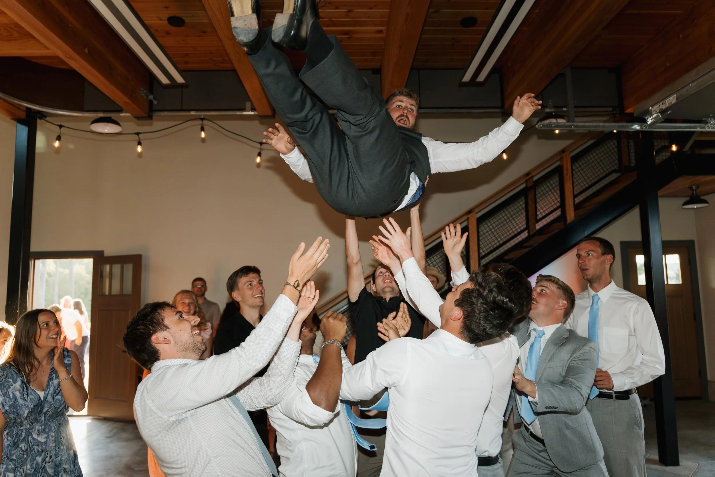 Group of people at a celebration lifting a man in a suit into the air indoors.
