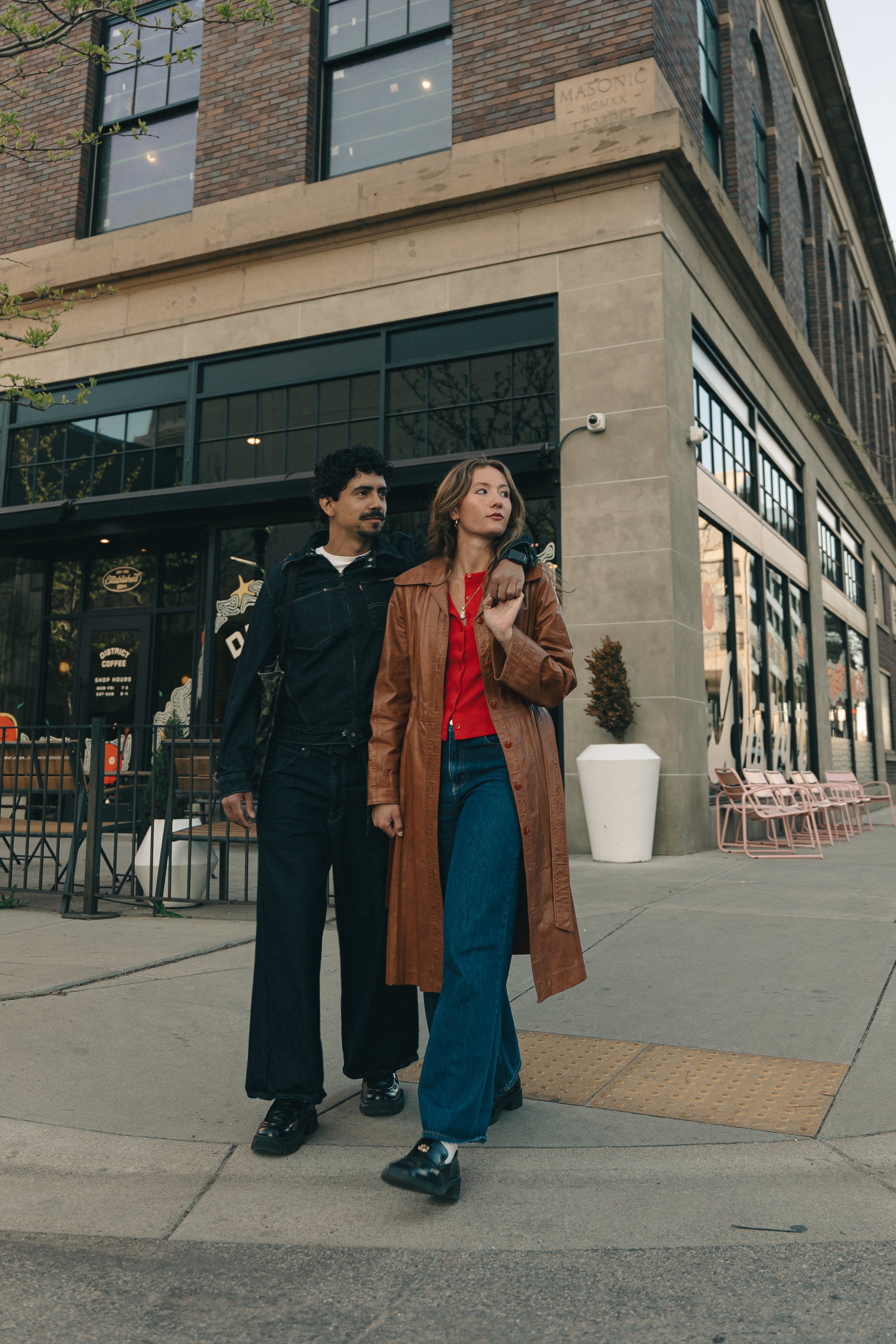 A man and woman walking on city sidewalk in front of a coffee shop, the man with his arm around the woman's shoulder, both dressed casually in jackets and jeans.
