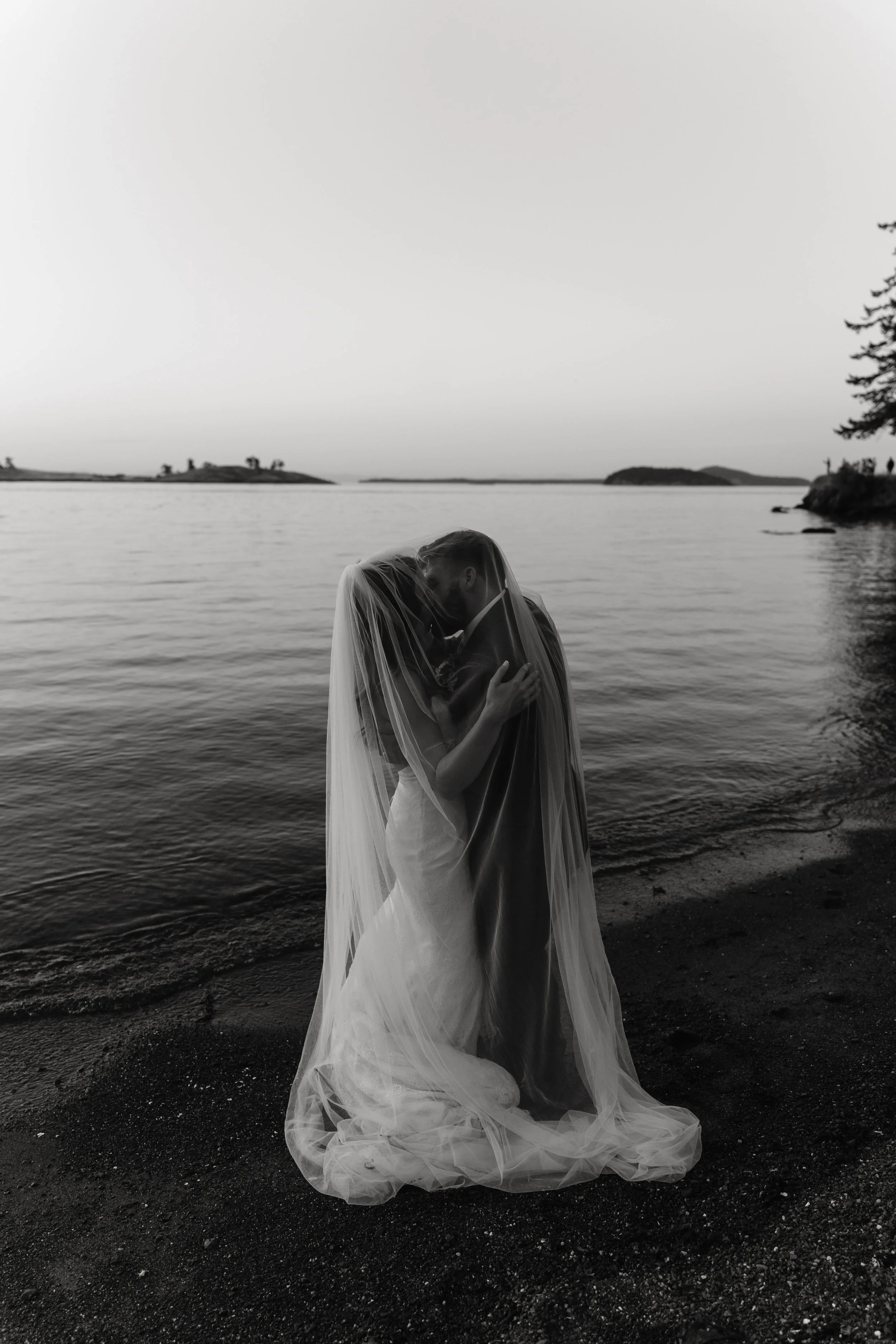 A bride and groom kissing on a beach at sunset, with a calm body of water and distant islands in the background, dressed in wedding attire, with the bride in a veil and gown.