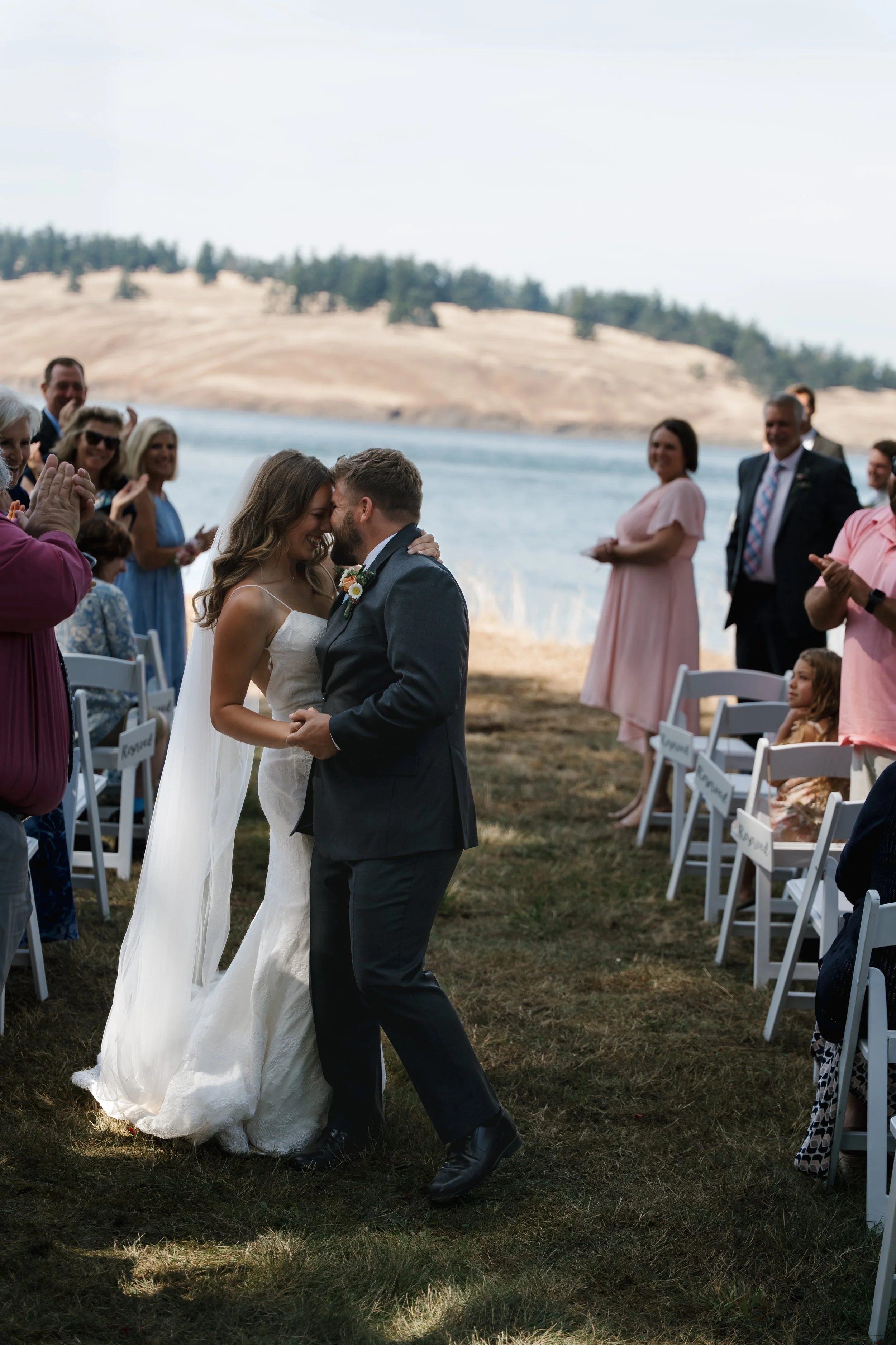 A newlywed couple kisses during an outdoor wedding by a lake, with guests watching and smiling.