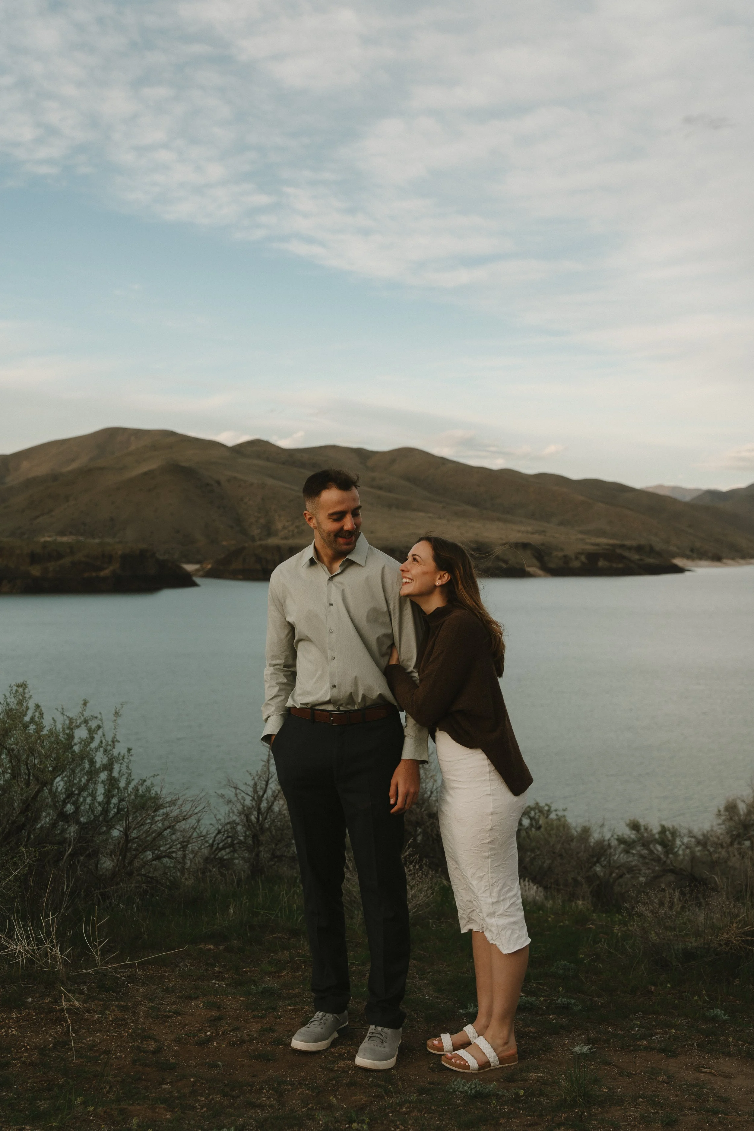 A couple standing near a body of water with mountains in the background, smiling at each other.