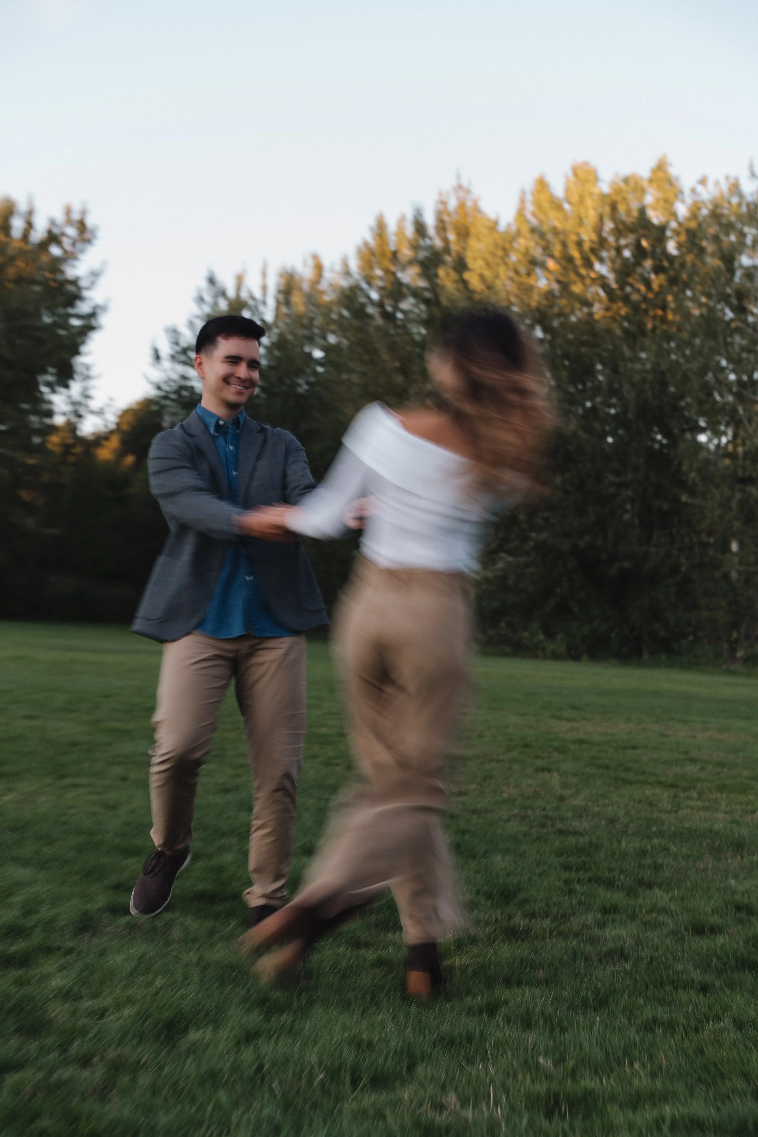 A man and woman holding hands, dancing on a grassy field with trees in the background. The woman appears blurred, indicating movement.