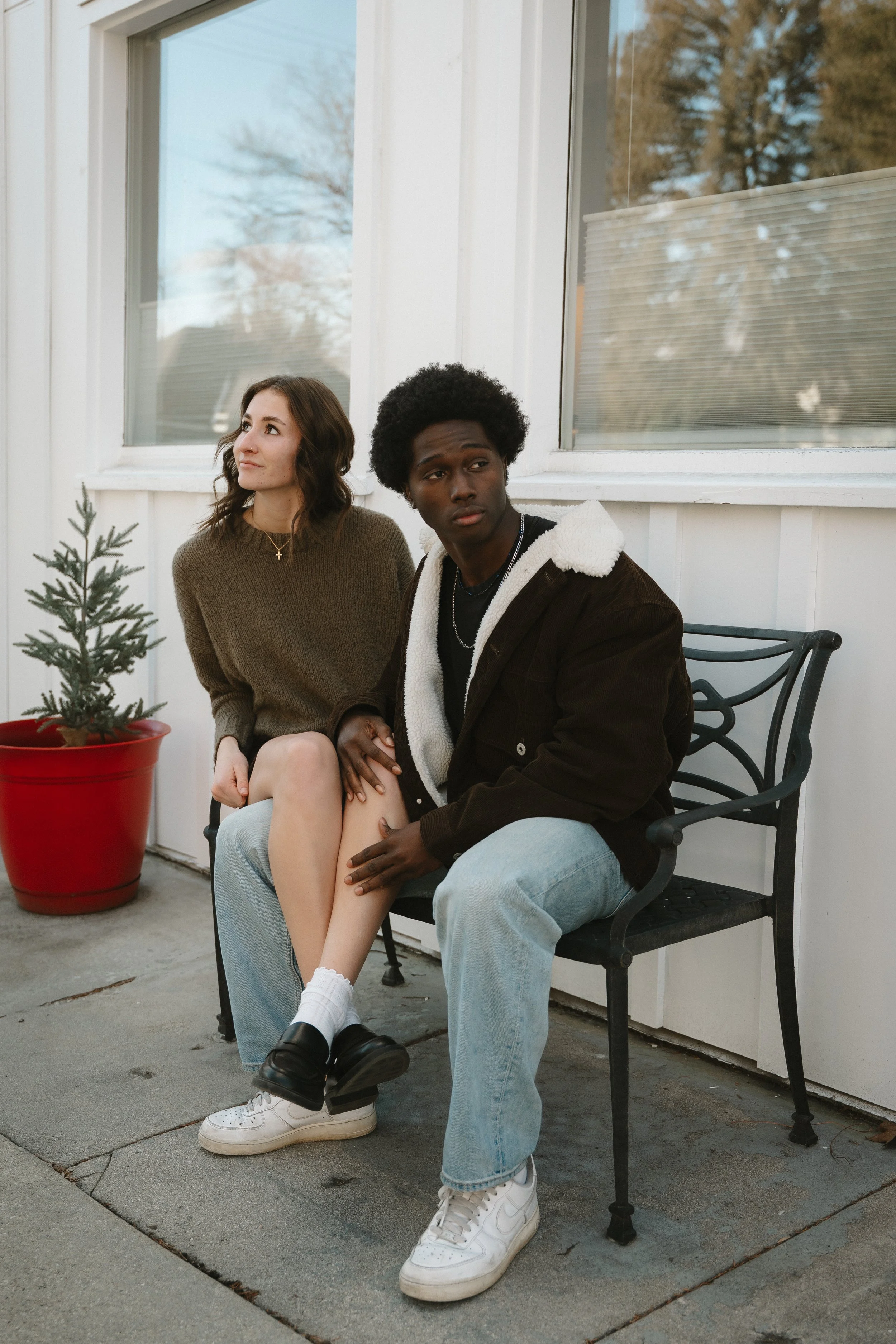 Two young people sitting on a bench outside near a white building and window, with a small potted Christmas tree nearby, during daytime.