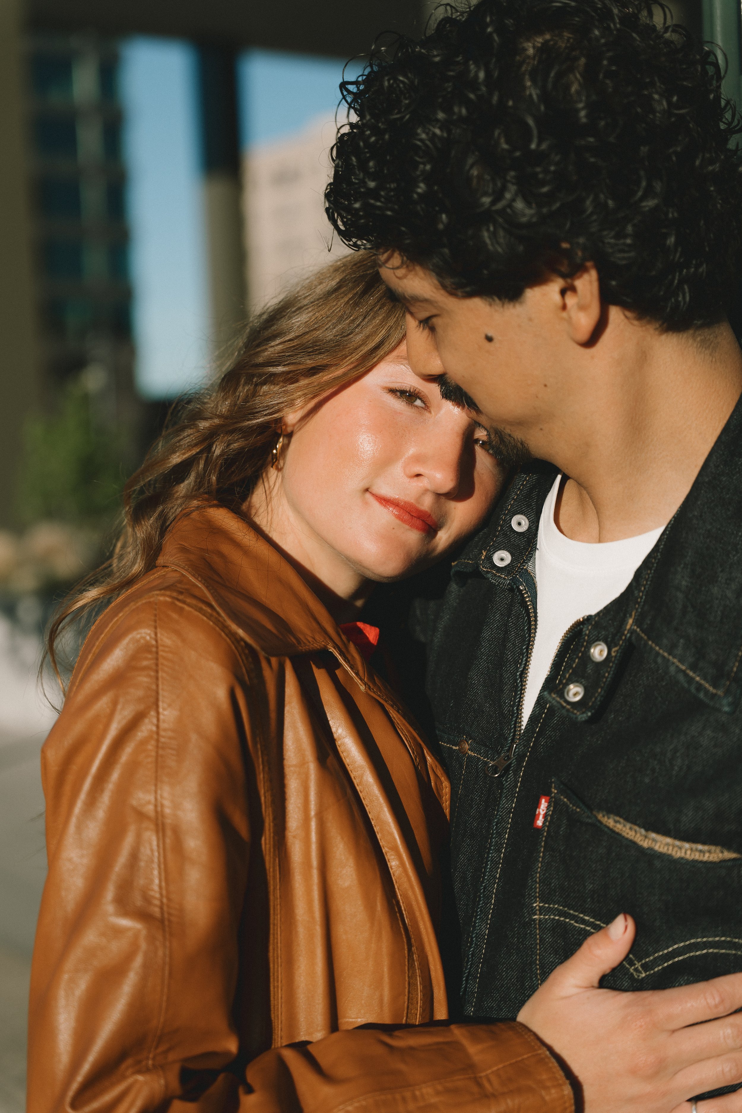 A young couple embracing outdoors in sunlight, with city buildings in the background. The woman has wavy light brown hair and is wearing a brown leather jacket, smiling softly. The man has curly dark hair, a mustache, and is wearing a dark denim jack