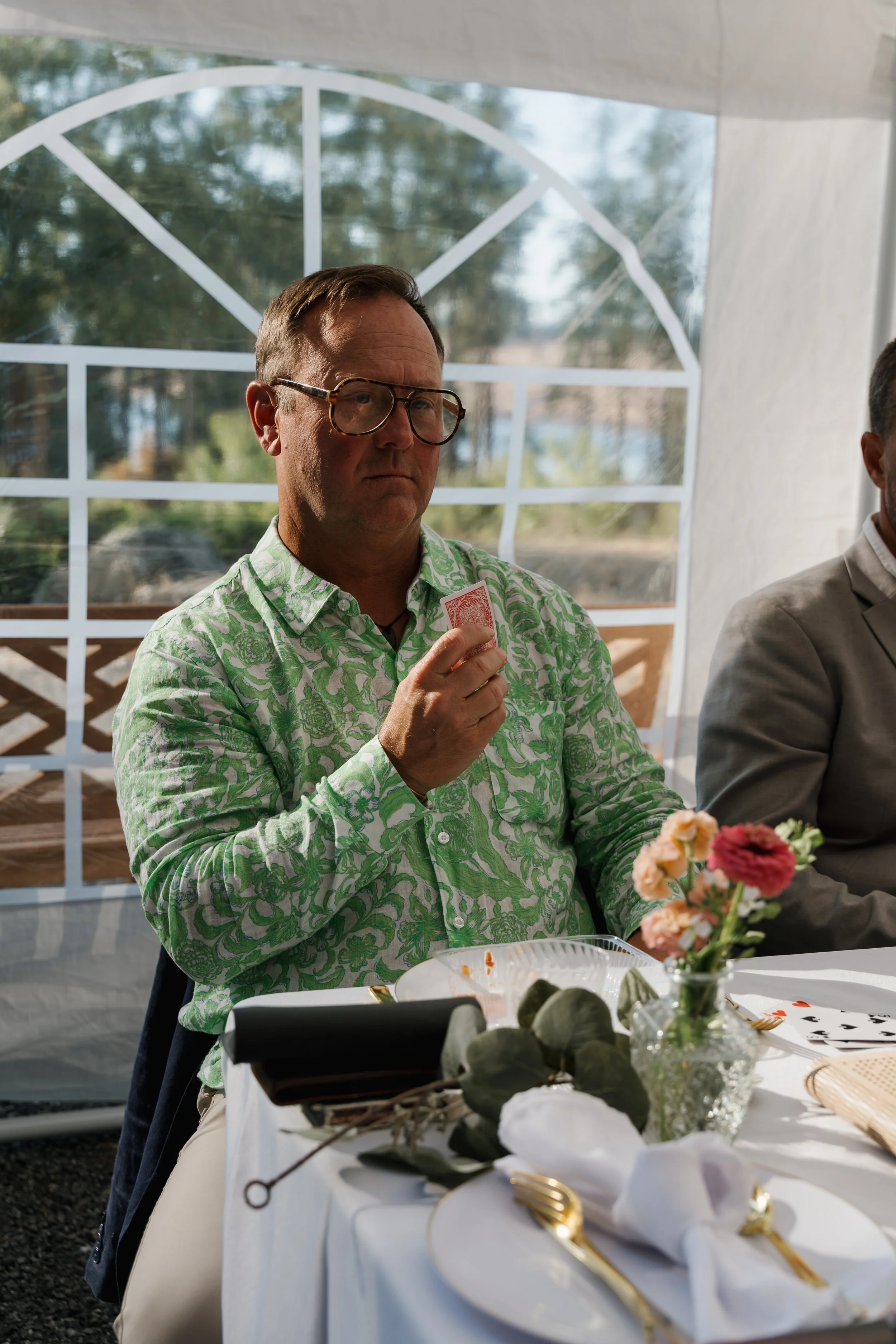 A man wearing glasses and a green floral shirt holds a playing card, sitting at a table decorated with flowers and plates during a gathering.